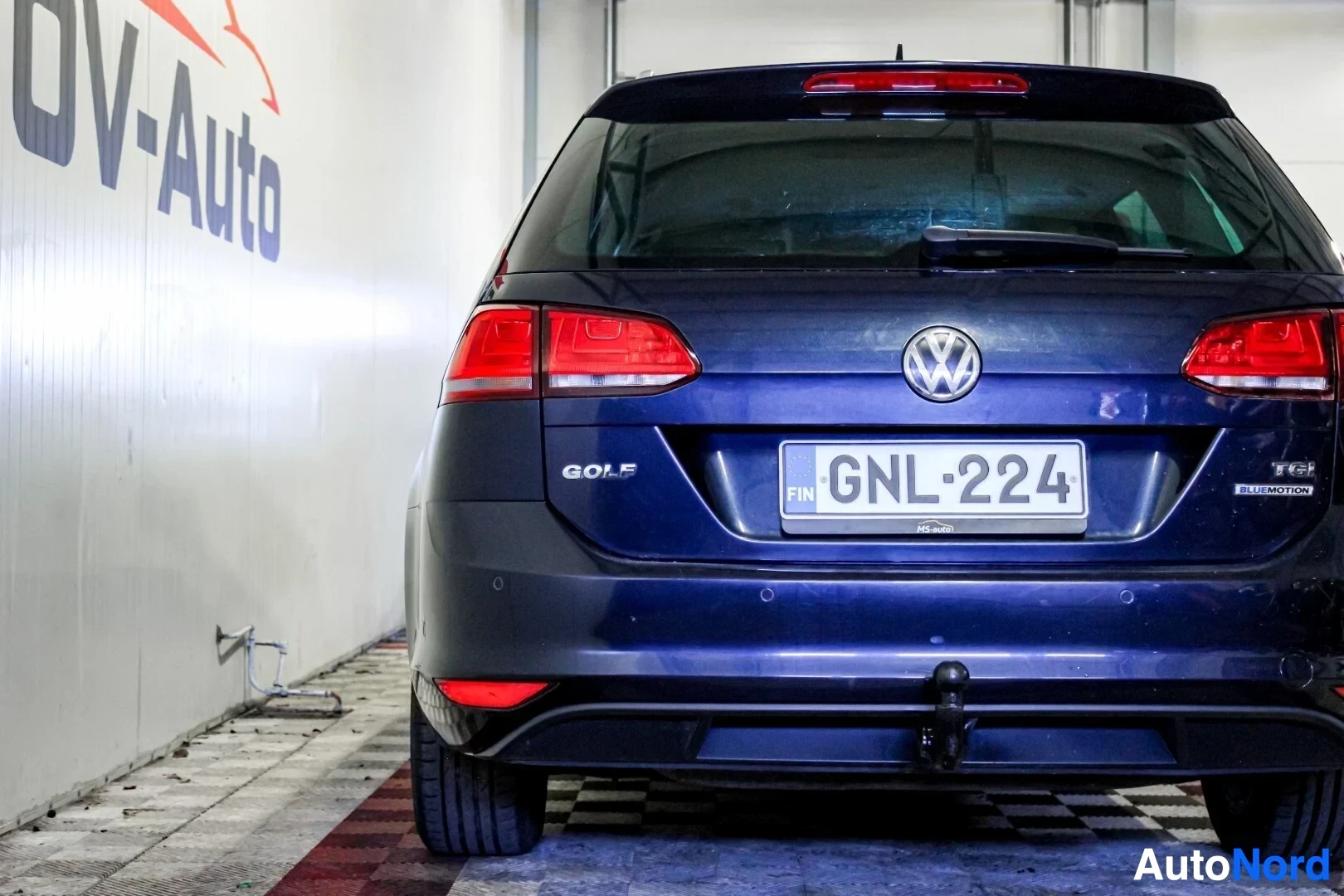 Takaa: Tumman farmariauton takaosa suoraan takaa kuvattuna autohallissa. Autossa on rekisterikilpi ja vetokoukku