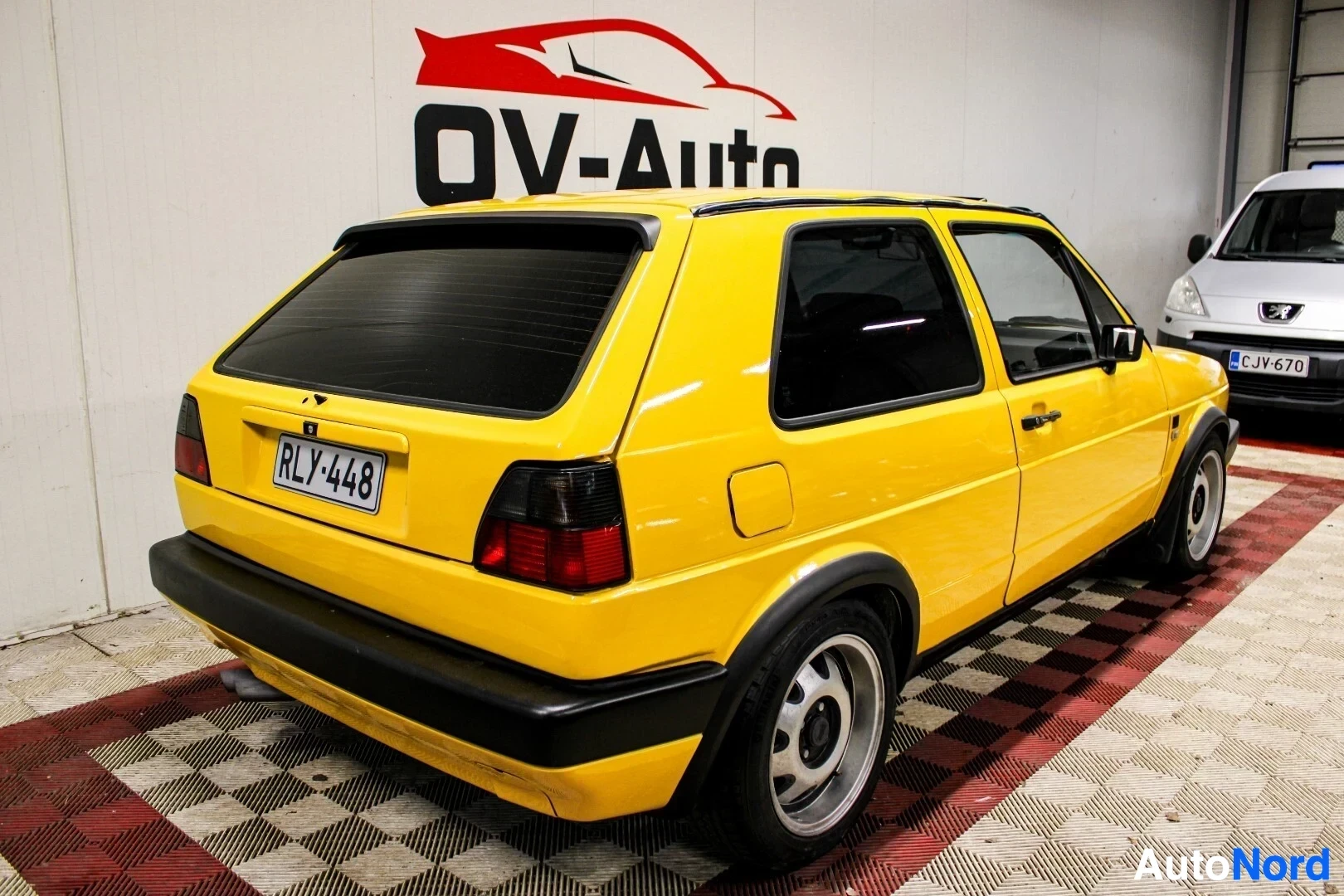 Takaviisto: Keltainen kaksiovinen hatchback, jossa on mustat puskurit ja takaspoileri, kuvattuna takaviistosta. Autossa on rekisterikilpi "". Tummaksi sävytetty taka