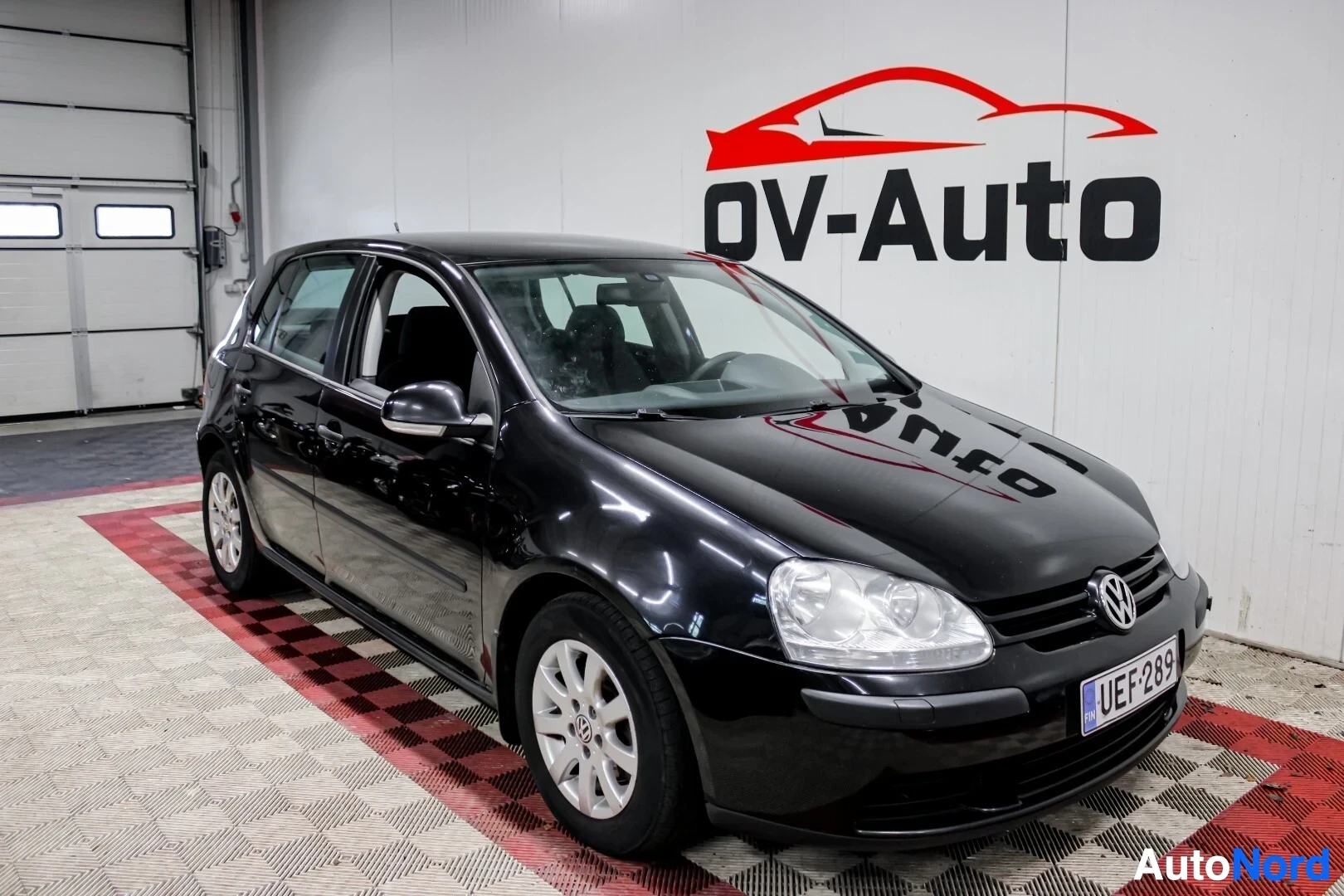 Etuviisto, nokka oikealle: Musta Volkswagen Golf viistosti edestä oikealta kuvattuna parkkihallissa. Autossa on hopeanväriset vanteet. Takana valkoisella seinällä on punamusta OV-A