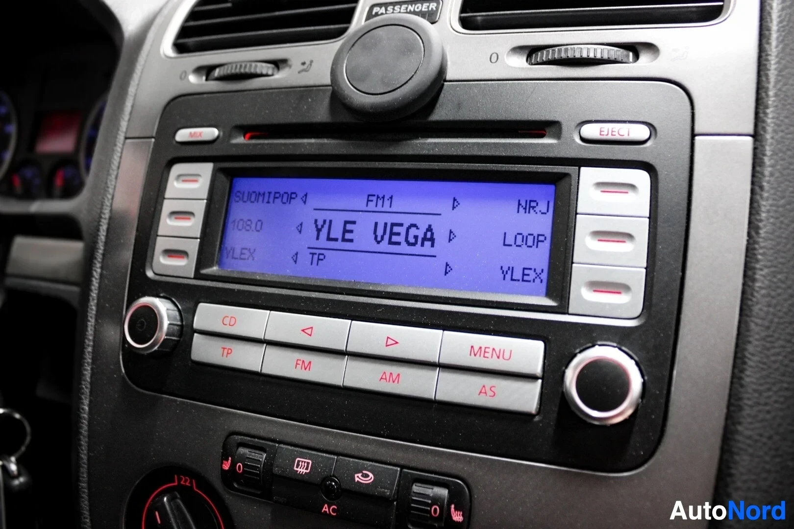Yksityiskohta: Lähikuva auton autoradion ja ilmastoinnin säätimistä. Radion näytöllä lukee "SUOMIPOP", "FM1", "NRJ", "108.0", "YLE VEGA", "LOOP", "YLEX" ja "TP".