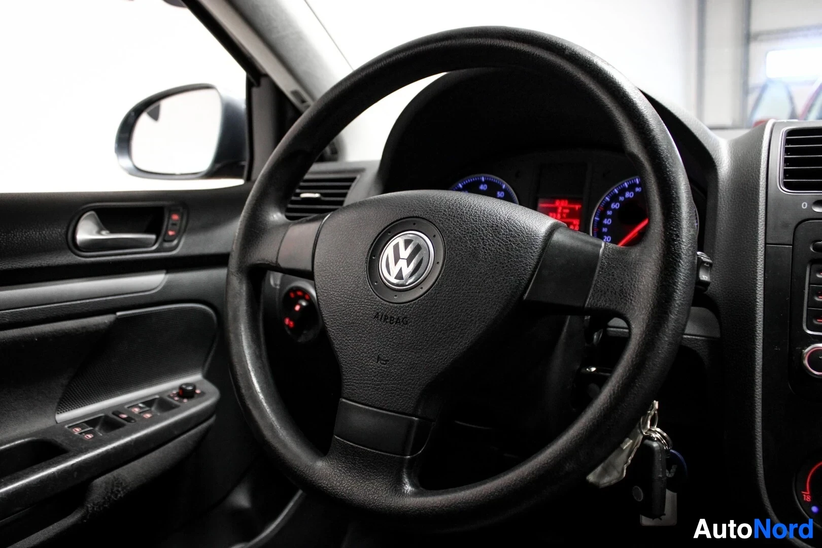 Ohjaamo: Sisäkuva auton ohjaamosta, jossa näkyvät ratti Volkswagen-logolla, mittaristo ja oven kyynärnoja ikkunan- ja peiliensäätimillä. Avaimet ovat virtalukossa.