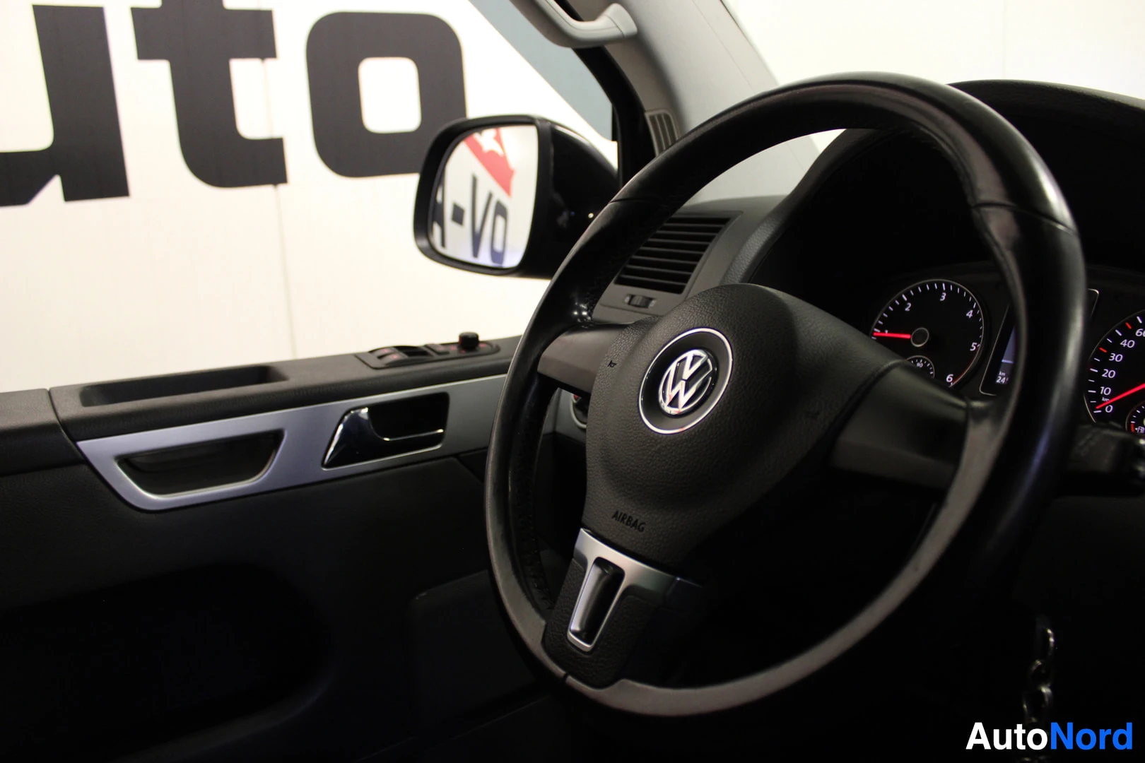 Ohjaamo: Lähikuva ohjaamon vasemmasta puolesta, jossa näkyy ohjauspyörä, jossa on Volkswagen-logo ja airbag-teksti, sekä osia kuljettajan ovesta ja mittaristosta. Tausta