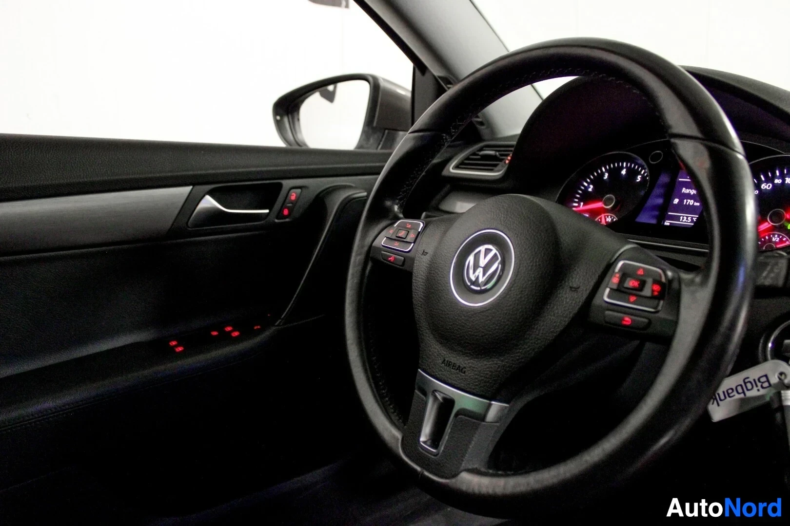 Ohjaamo: Kuljettajan oven sisäpaneeli ja ohjauspyörä, jossa on painikkeita ja Volkswagen-logo.