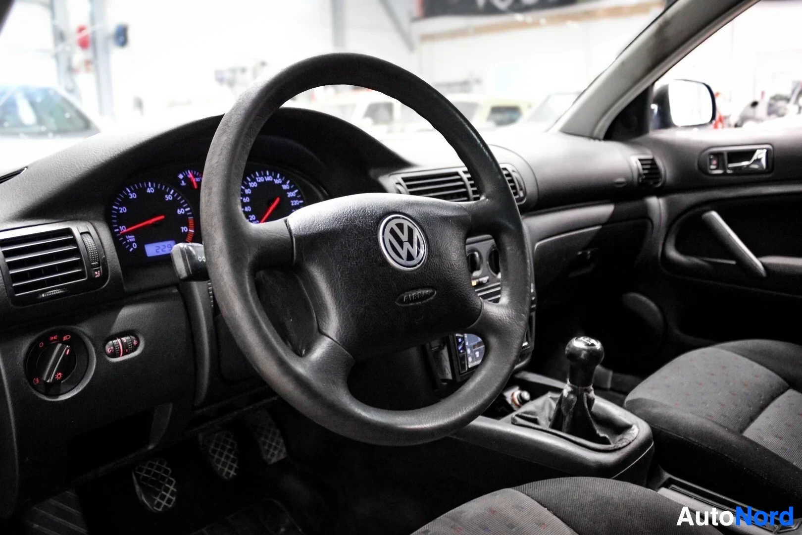 Ohjaamo: Musta ohjauspyörä, jossa VW-logo keskellä, mittaristo, keskikonsoli ja manuaalivaihteisto näkyvissä.