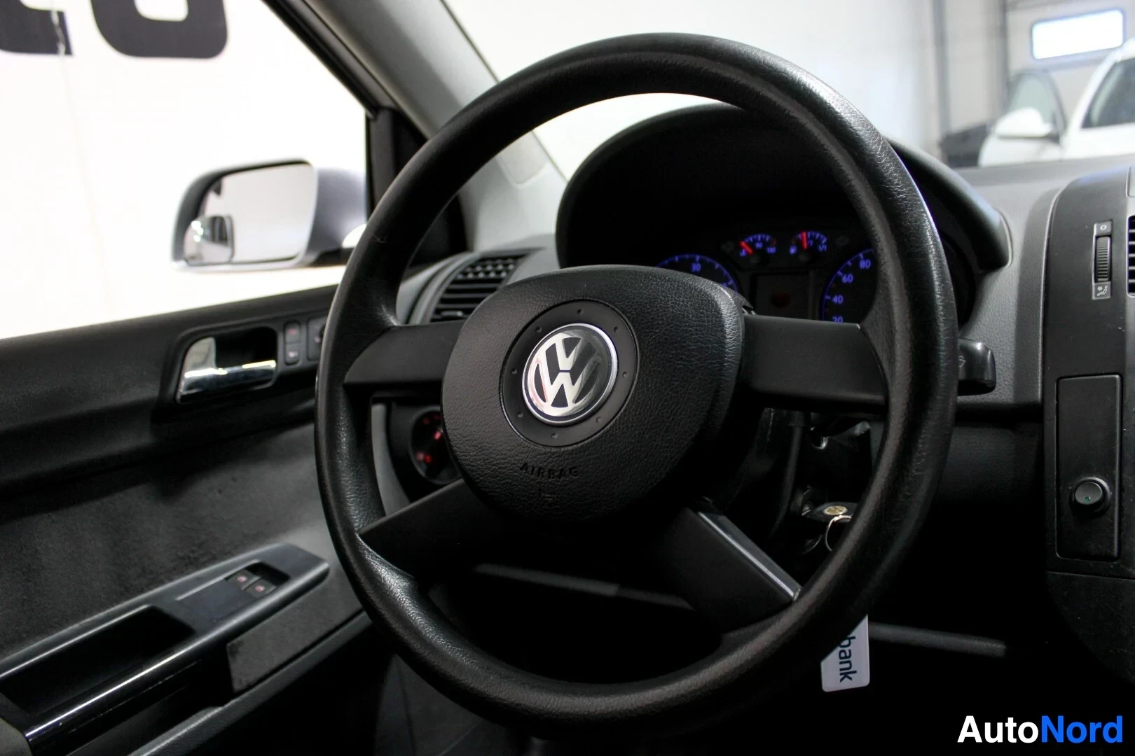 Yksityiskohta: Ohjauspyörä, jossa on Volkswagen-logo ja "AIRBAG"-teksti. Ovipaneeli ja sivupeili näkyvät vasemmalla.