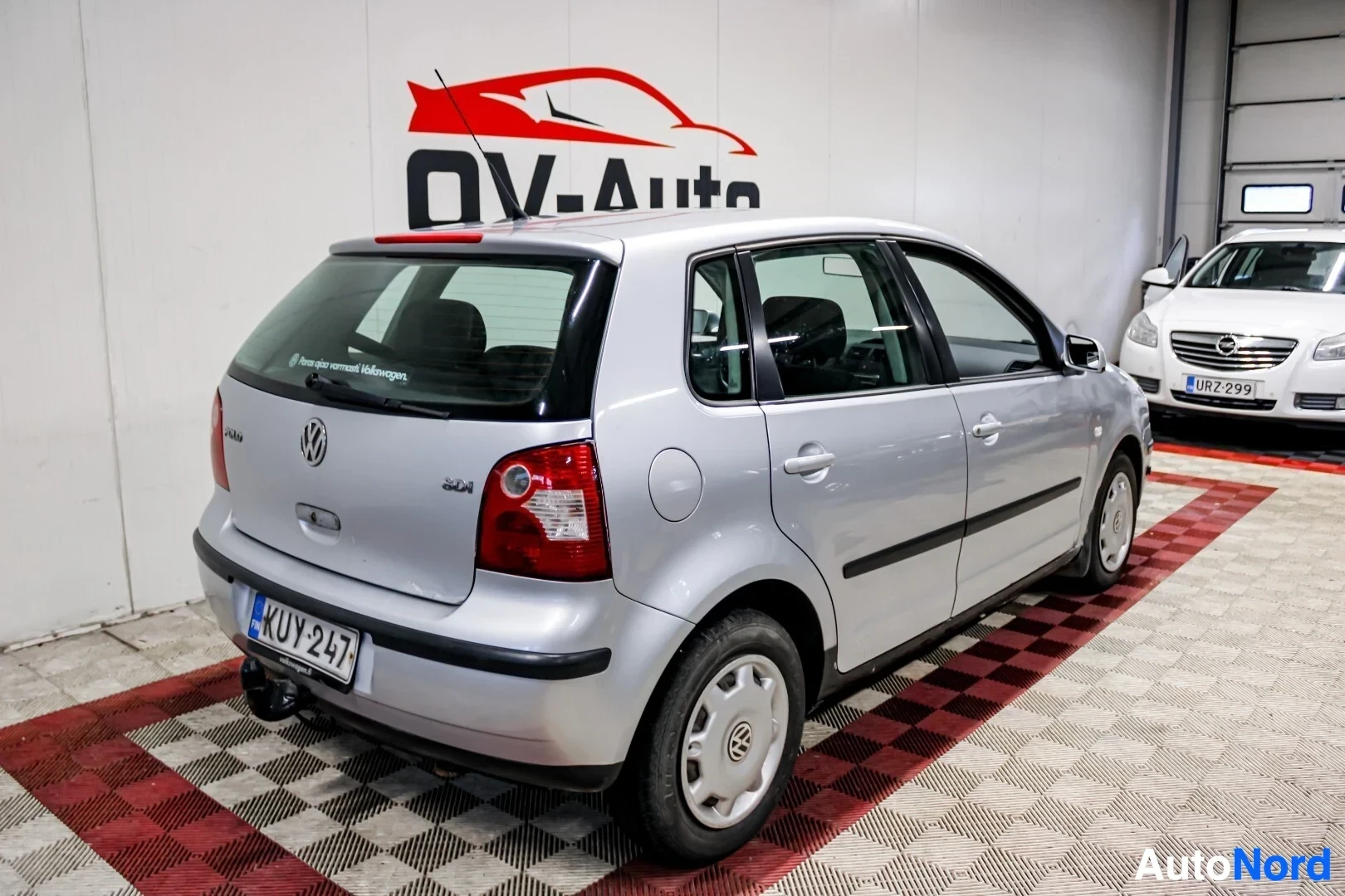 Takaviisto: Hopeanvärinen Volkswagen Polo -henkilöauto pysäköitynä valkoisen seinän edessä. Auto on kuvattu takaapäin viistosti oikealta. Autossa on vetokoukku, takaluukuss