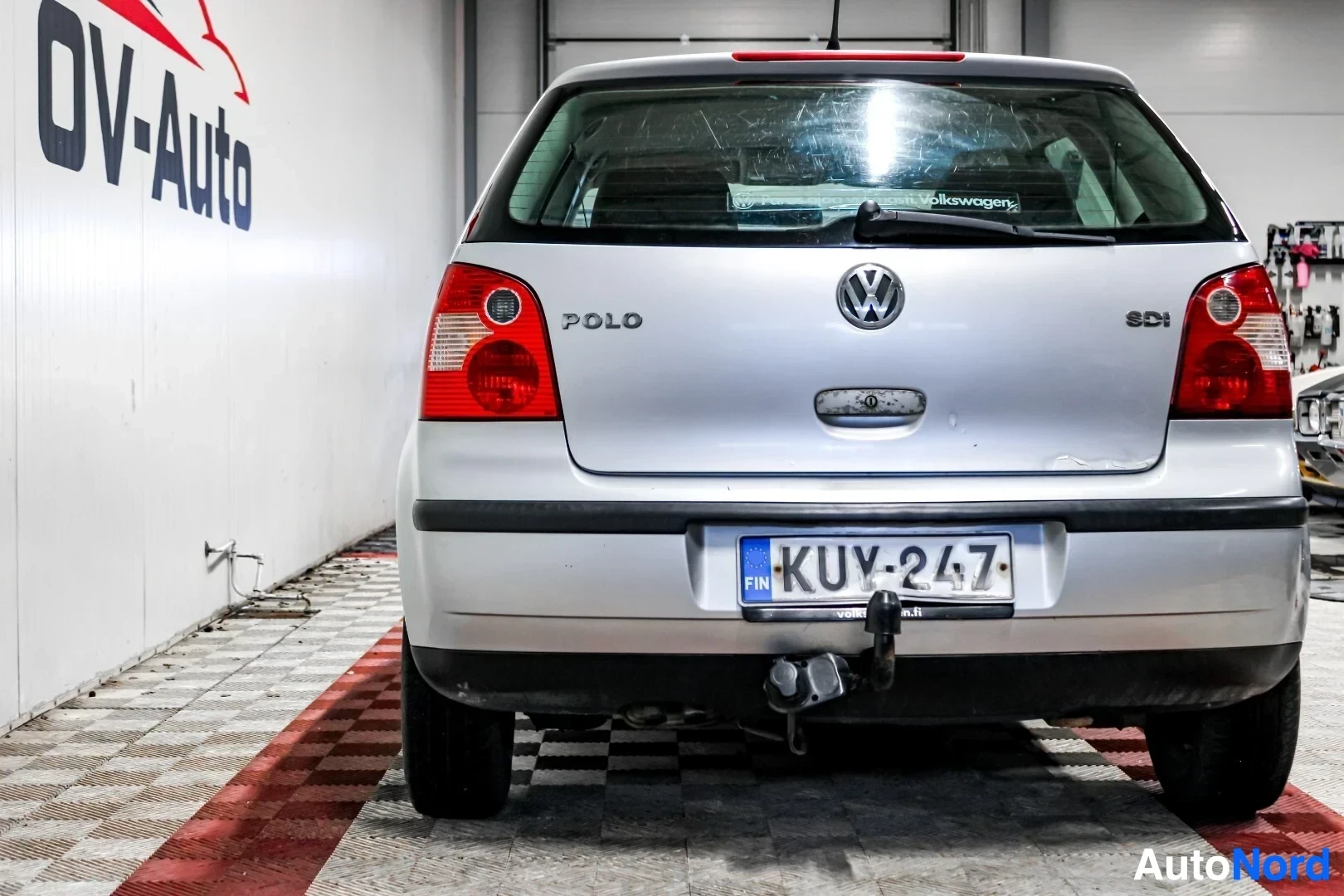 Takaa: Hopeanvärinen Volkswagen Polo -henkilöauto pysäköitynä valkoisen seinän edessä. Auto on kuvattu takaa. Autossa on vetokoukku, takaluukussa on Volkswagen-logo ja