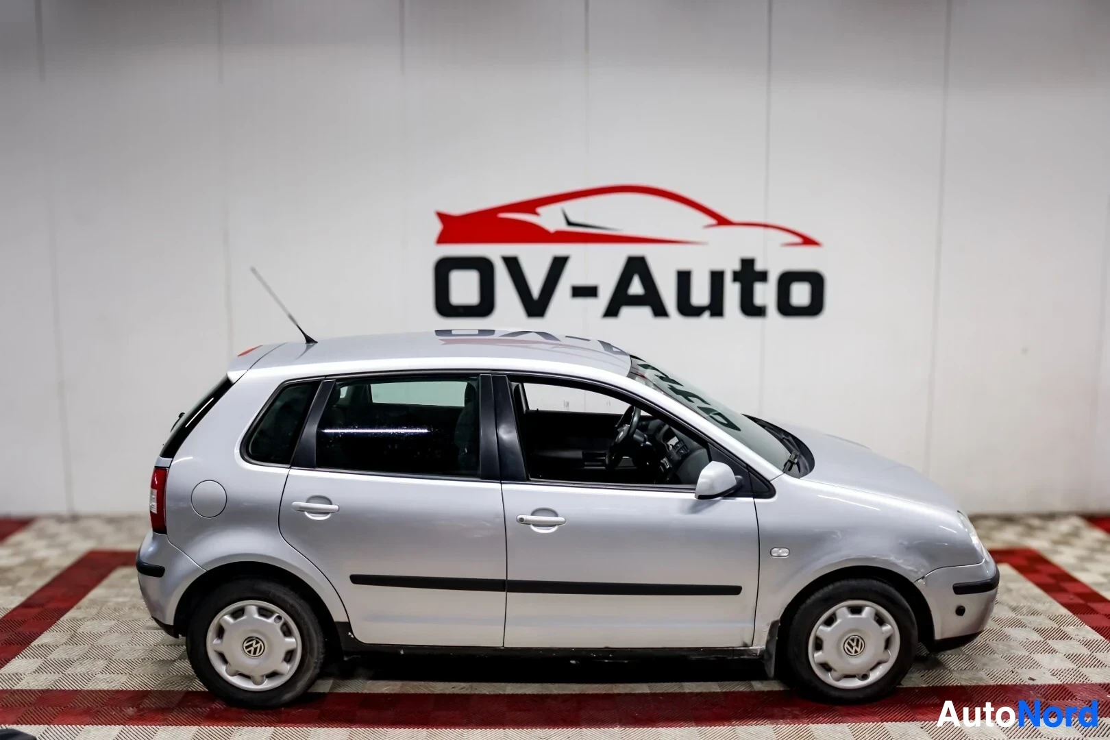 Sivulta: Hopeanvärinen Volkswagen Polo -henkilöauto pysäköitynä valkoisen OV-Auto-logon edessä. Auto on kuvattu kyljittäin vasemmalta puolelta. Autossa on viisipuolaiset
