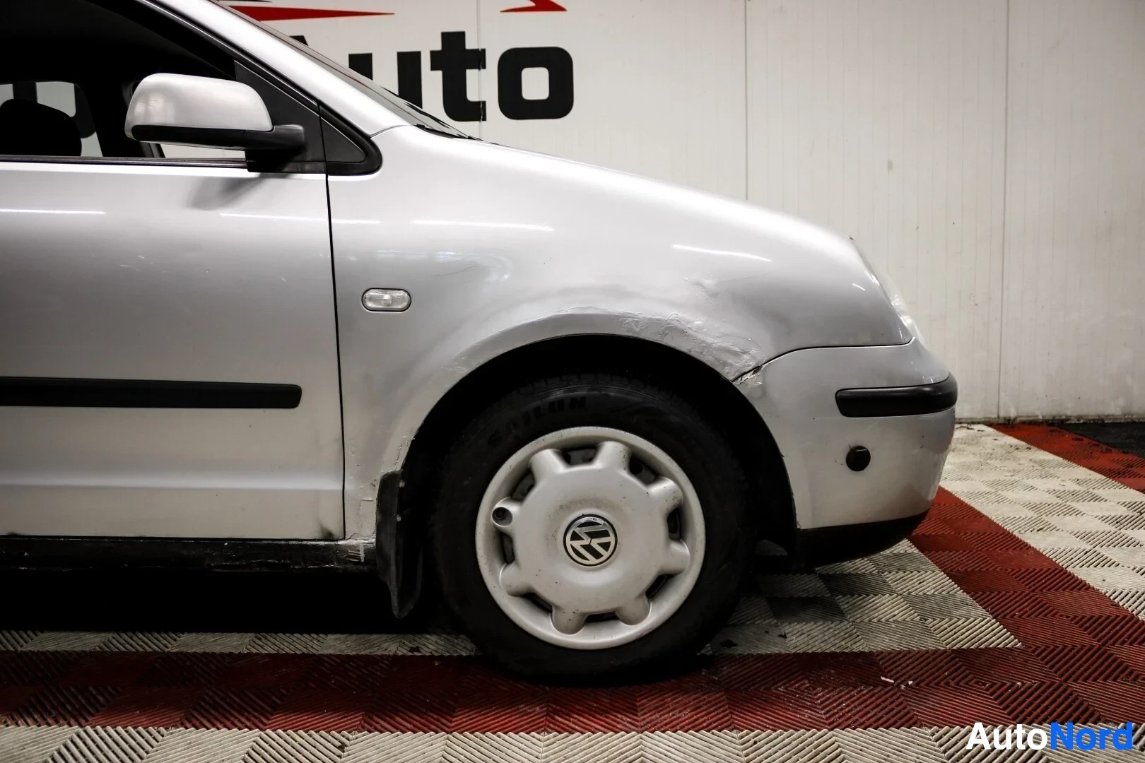 Yksityiskohta: Lähikuva hopeanvärisen auton etupyörästä ja etulokasuojasta. Lokasuojassa ja etupuskurissa näkyy kulumaa ja vaurioita. Pyörässä on peltivanne ja Volkswagen-logo