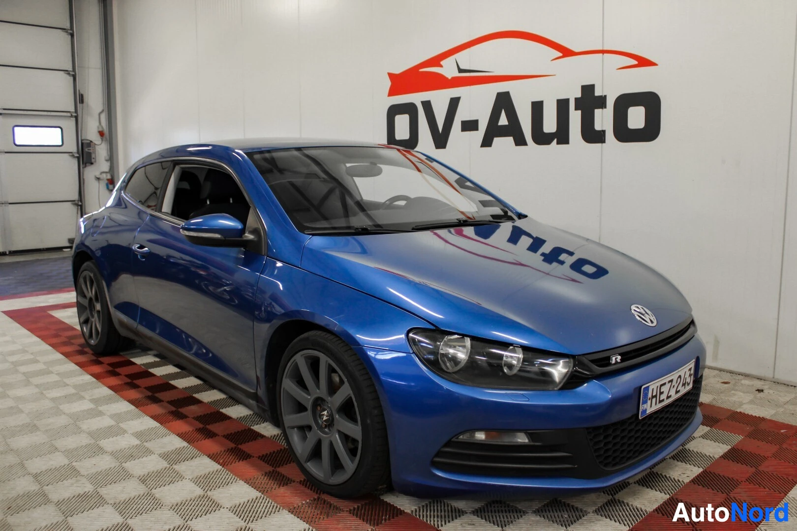 Etuviisto, nokka oikealle: Sinisestä autosta otettu kuva edestä ja sivulta sisältä. Auto on pysäköity sisätilaan ruudulliselle lattialle, ja taustalla on OV-Auton logo.