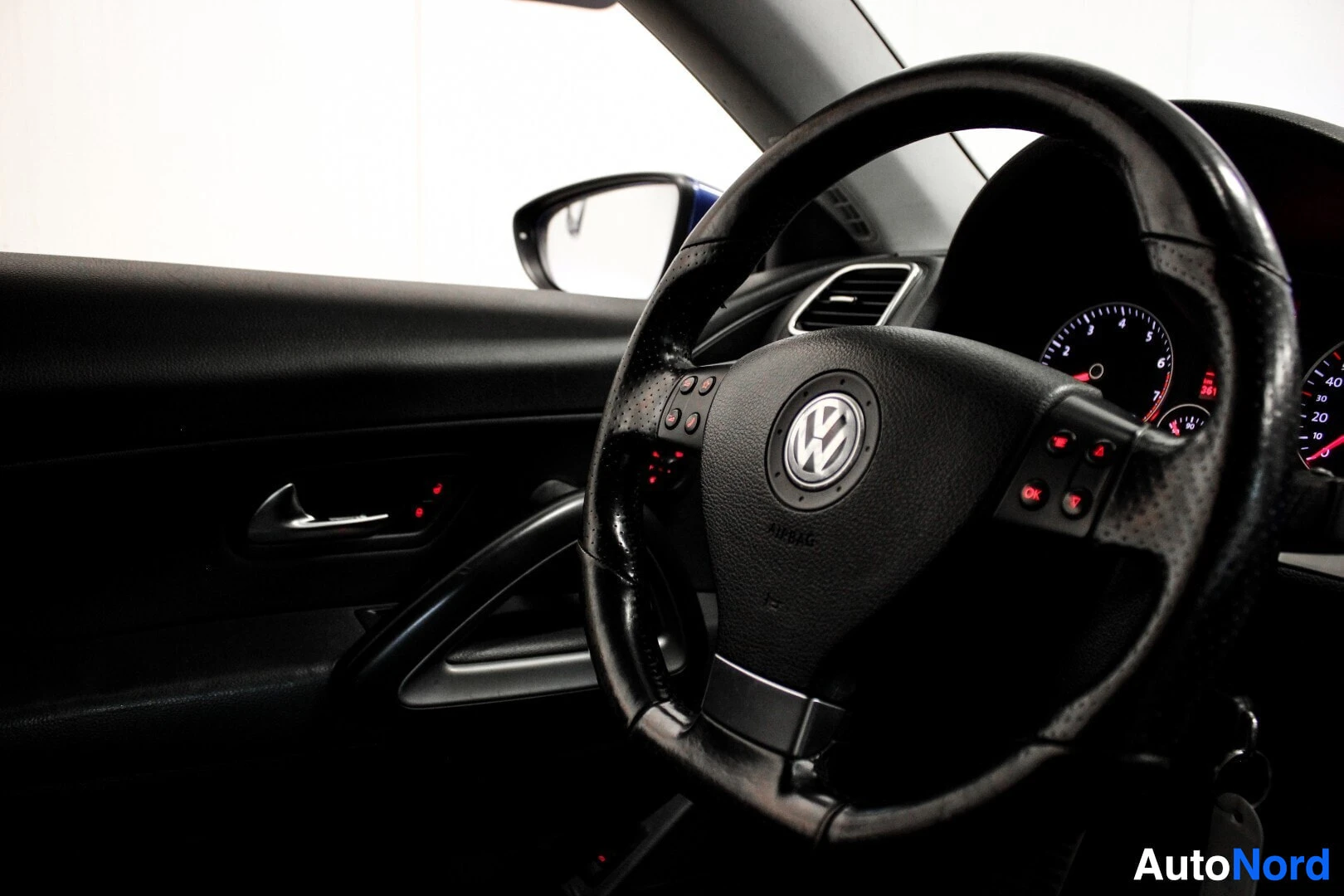Ohjaamo: Musta nahkainen monitoimiohjauspyörä Volkswagen-logolla, auton sisustus ja ulkopeili heijastuksineen.