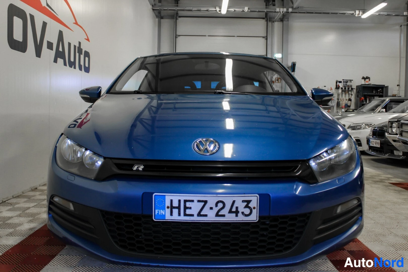 Edestä: Sinisestä autosta otettu etukuva sisältä. Auto on pysäköity sisätilaan ruudulliselle lattialle, ja taustalla on OV-Auton logo. Auton rekisterikilpi on