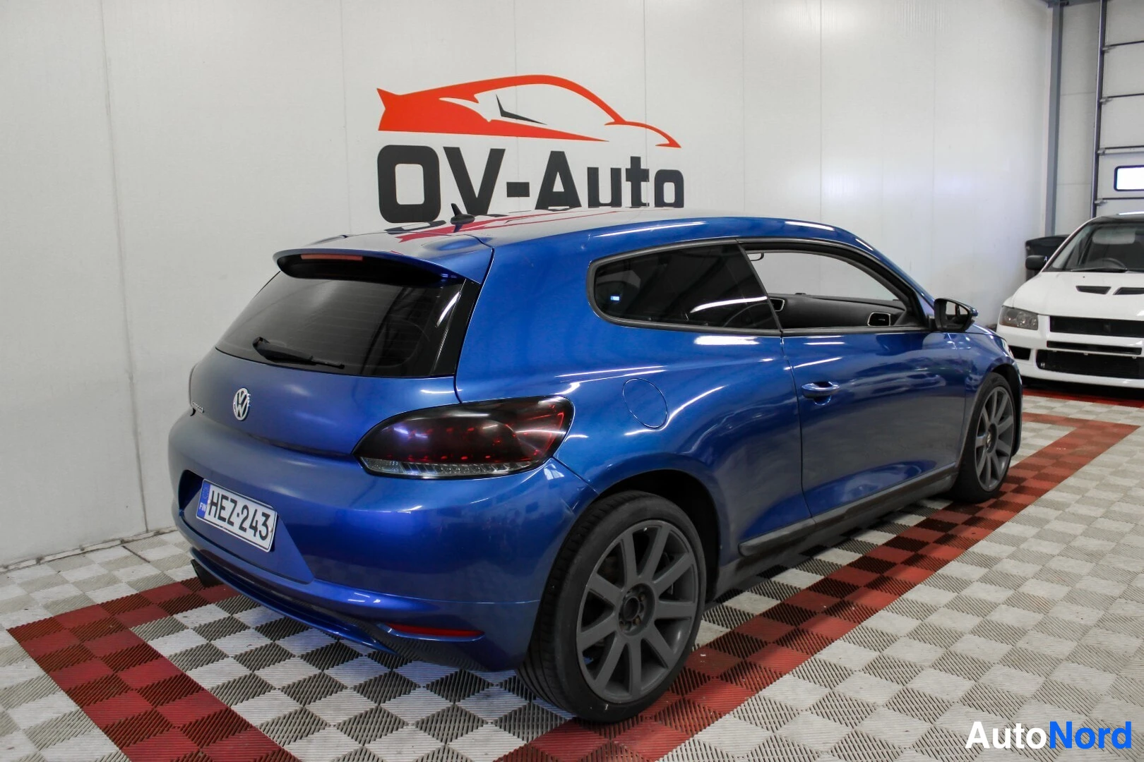 Takaviisto: Sinisestä autosta otettu takaviistokuva sisältä. Auto on pysäköity sisätilaan ruudulliselle lattialle, ja taustalla on OV-Auton logo. Oikealla taustalla näkyy t