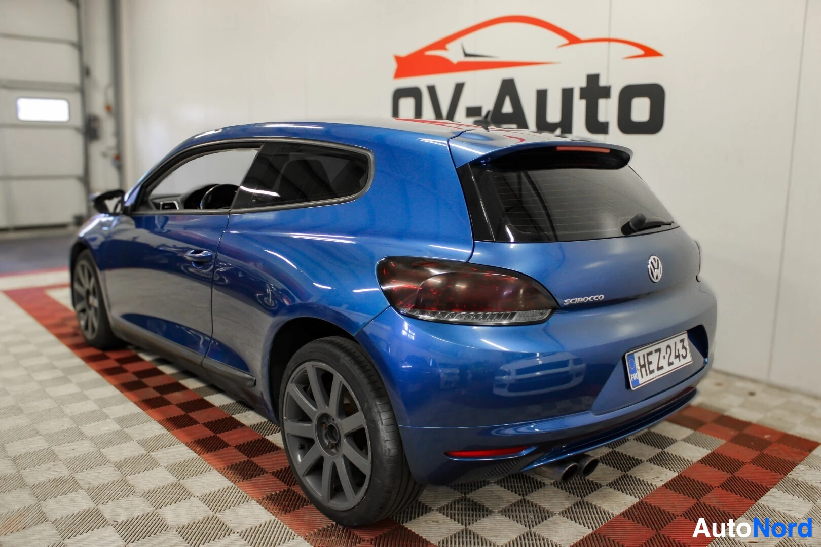 Takaviisto: Sivuttain takaoikealta kuvattu sininen coupé-auto pysäköitynä valkoiselle taustalle, jossa on autoliikkeen logo.