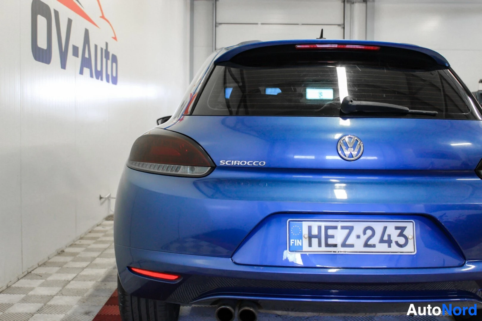 Takaa: Lähikuva sinisen coupé-auton takaosasta, jossa näkyy takarekisterikilpi, tavaratilan kansi ja takavalot. Taustalla autoliikkeen logo.