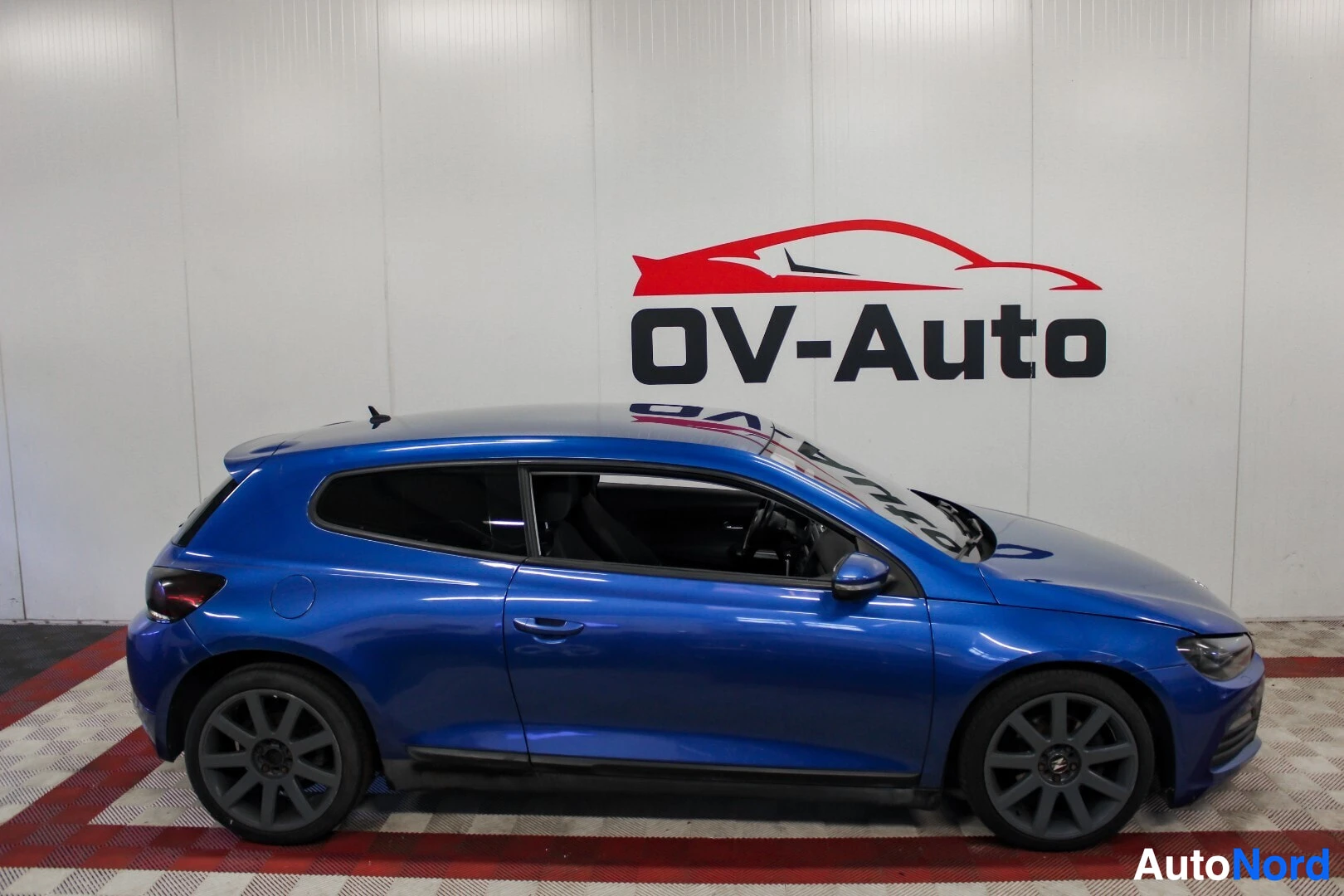 Sivulta: Sininen coupé-auto kuvattuna sivusta, vasemmalta puolelta. Auto on pysäköity valkoiselle taustalle, jossa on autoliikkeen logo.