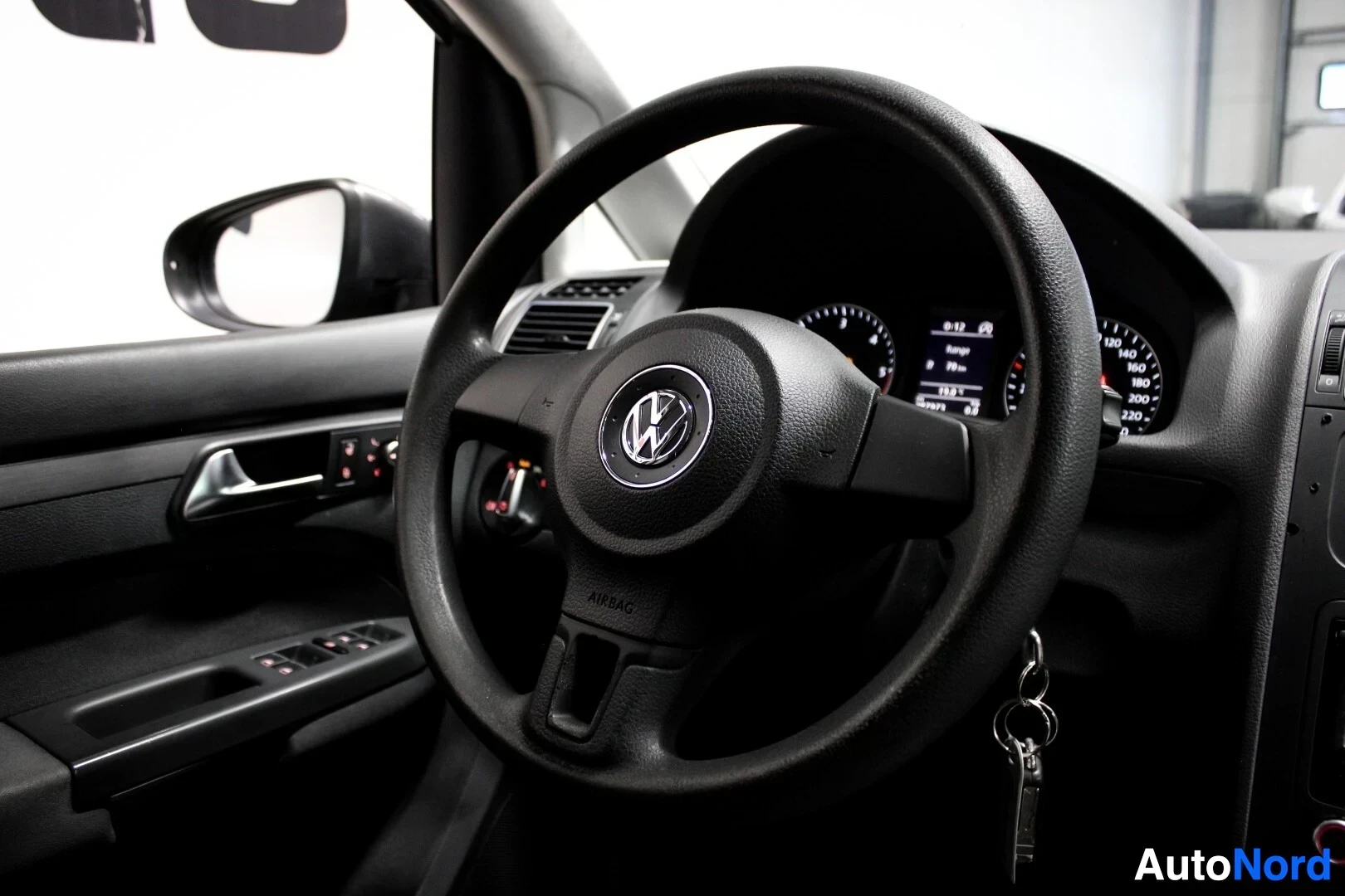 Ohjaamo: Ajoneuvon ohjaamo kuvattuna kuljettajan puolelta, keskittyen ohjauspyörään ja oven sisäpaneeliin. Ohjauspyörässä on VW-logo ja alareunassa teksti "AIRBAG". Oven
