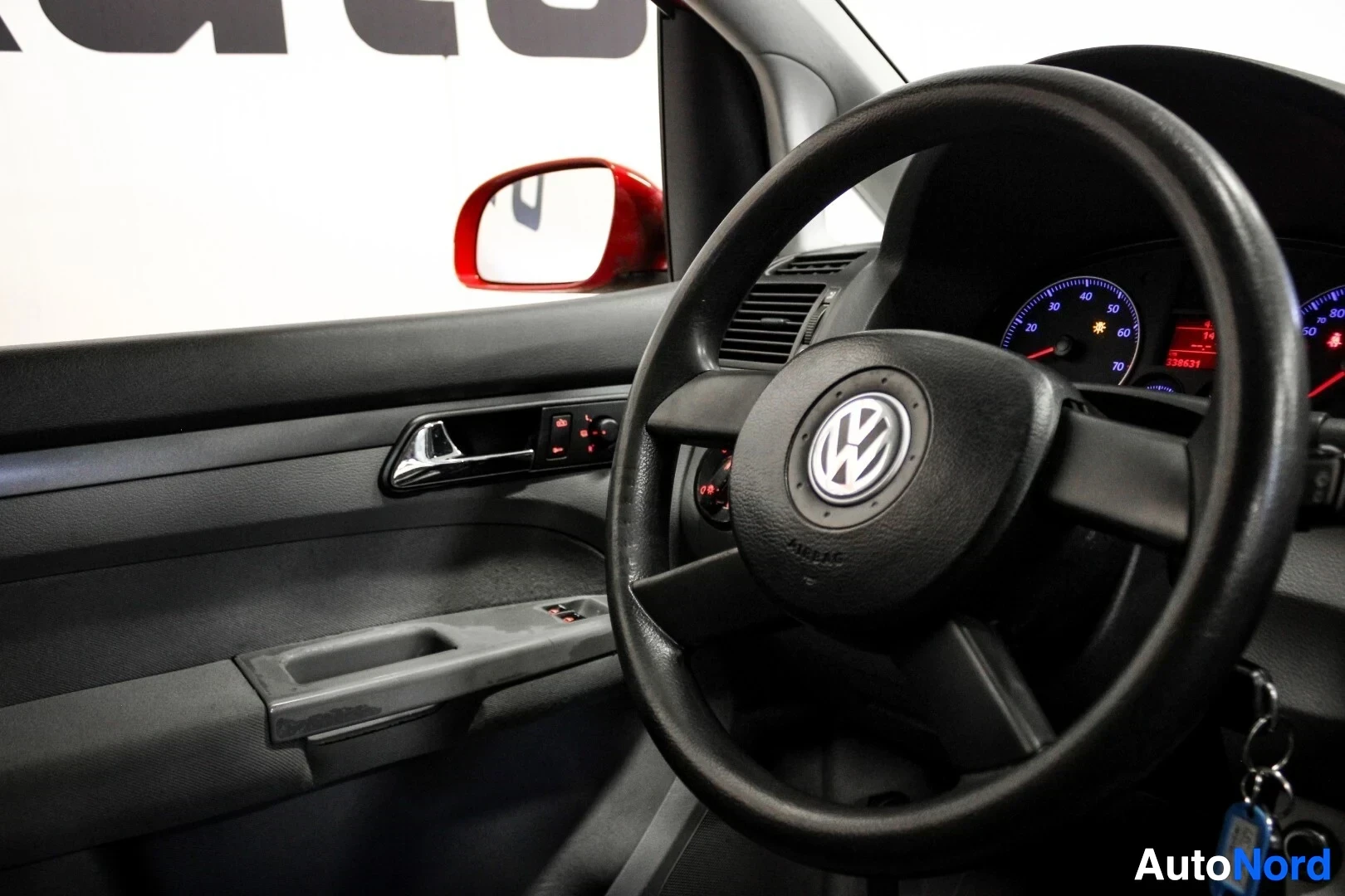 Ohjaamo: Ohjauspyörä Volkswagen-logolla, vasen etuovi avattuna ja punainen sivupeili näkyvissä.