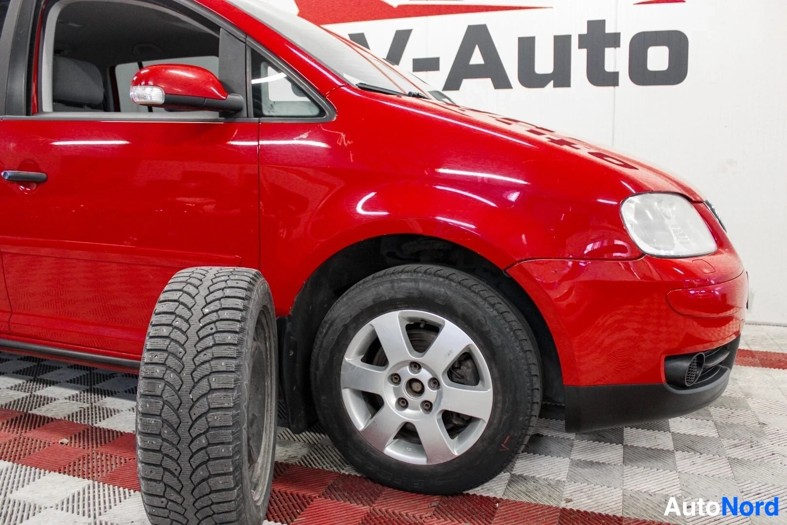 Etuviisto, nokka vasemmalle: Punaista henkilöautoa edestä oikealta puolelta, oikea eturengas ja sen vieressä erillinen nastoitettu talvirengas. Taustalla näkyy 'V-Auto' logo.