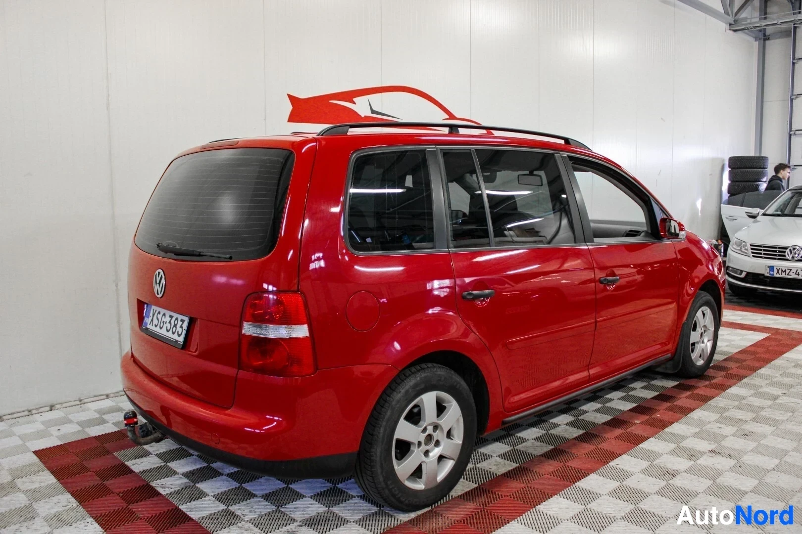 Takaviisto: Punainen henkilöauto, jossa on kattokaiteet ja vetokoukku, seisoo autonäyttelyn lattialla. Auton takaosa ja oikea kylki näkyvät kuvassa, vasen takarengas ja vet