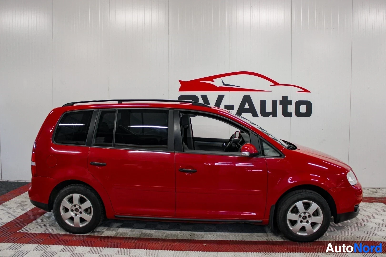 Sivulta: Punainen tila-auto, jossa on kattokaiteet ja viisipuolaiset kevytmetallivanteet, seisoo lattialla. Koko auton oikea kylki on näkyvissä.