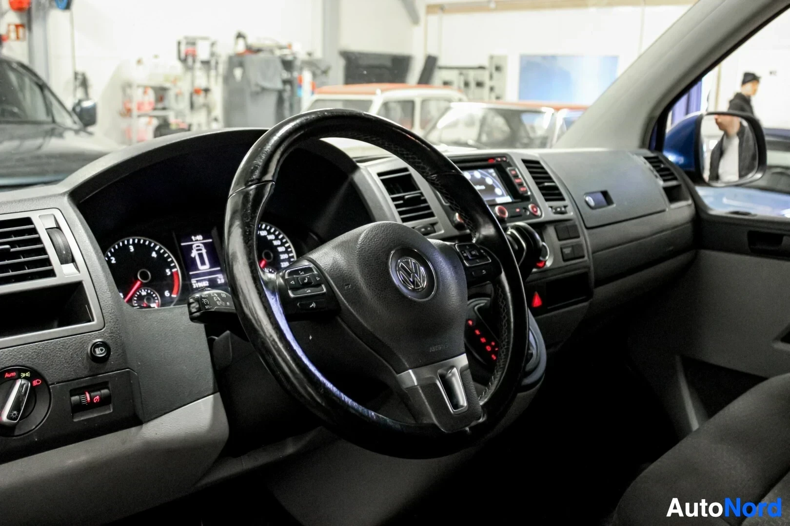 Ohjaamo: Kuvakulma auton matkustamoon kuljettajan puolelta, näyttäen ohjauspyörän ja kojelaudan. Ohjauspyörässä on multimediapainikkeita ja Volkswagenin logo. Keskikonso