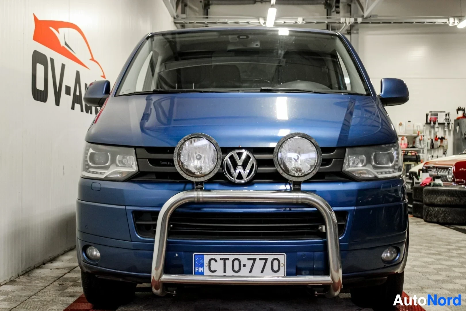 Edestä: Sinisen pakettiauton etuosa, jossa on rekisterikilpi "", keulaputket ja kaksi suurta pyöreää lisävaloa. Edessä on Volkswagenin logo