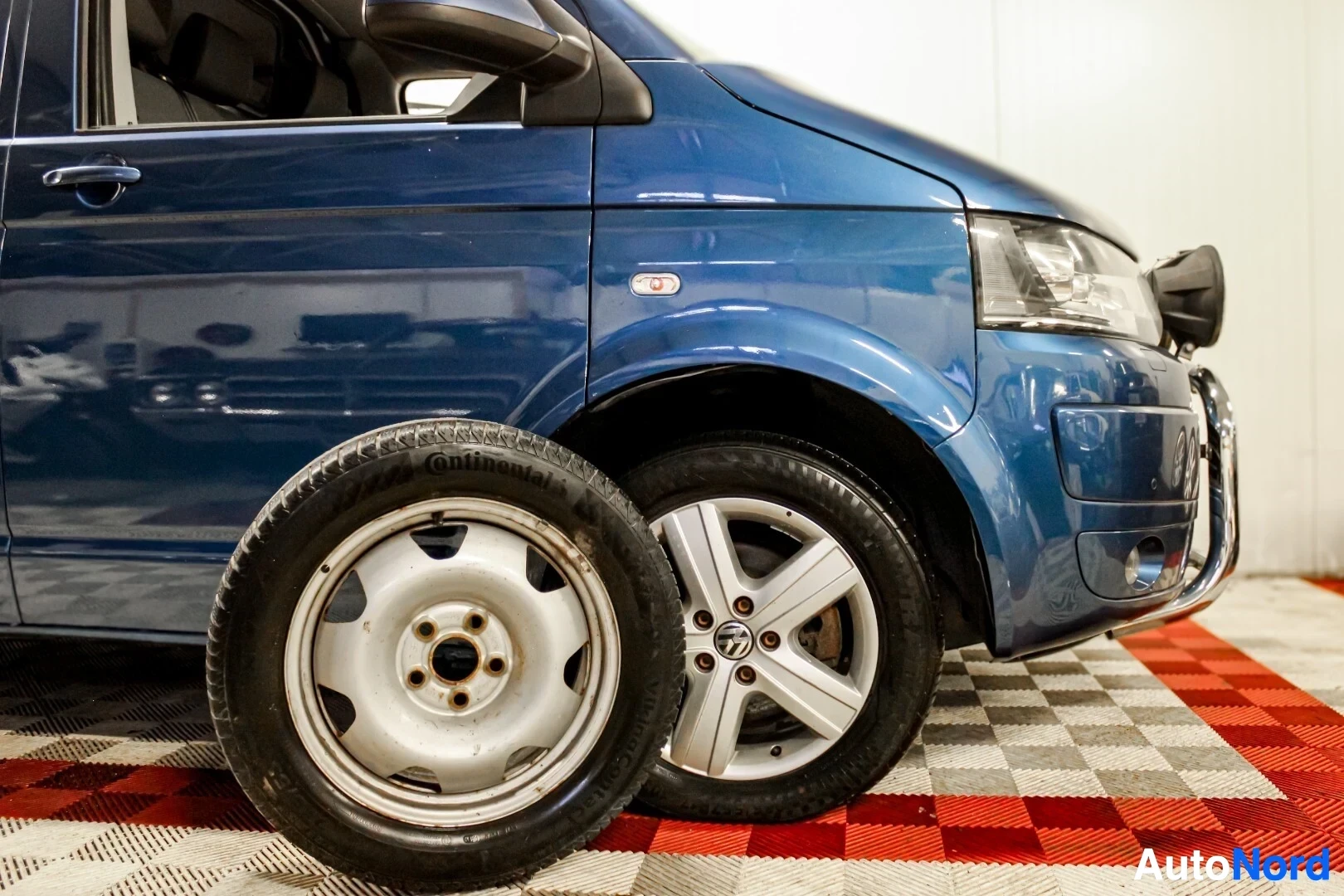 Yksityiskohta: Sinisen pakettiauton etupäästä otettu lähikuva. Kuvan etualalla näkyy kaksi auton rengasta: toinen on kiinni autossa ja siinä on kevytmetallivanne, toinen seiso