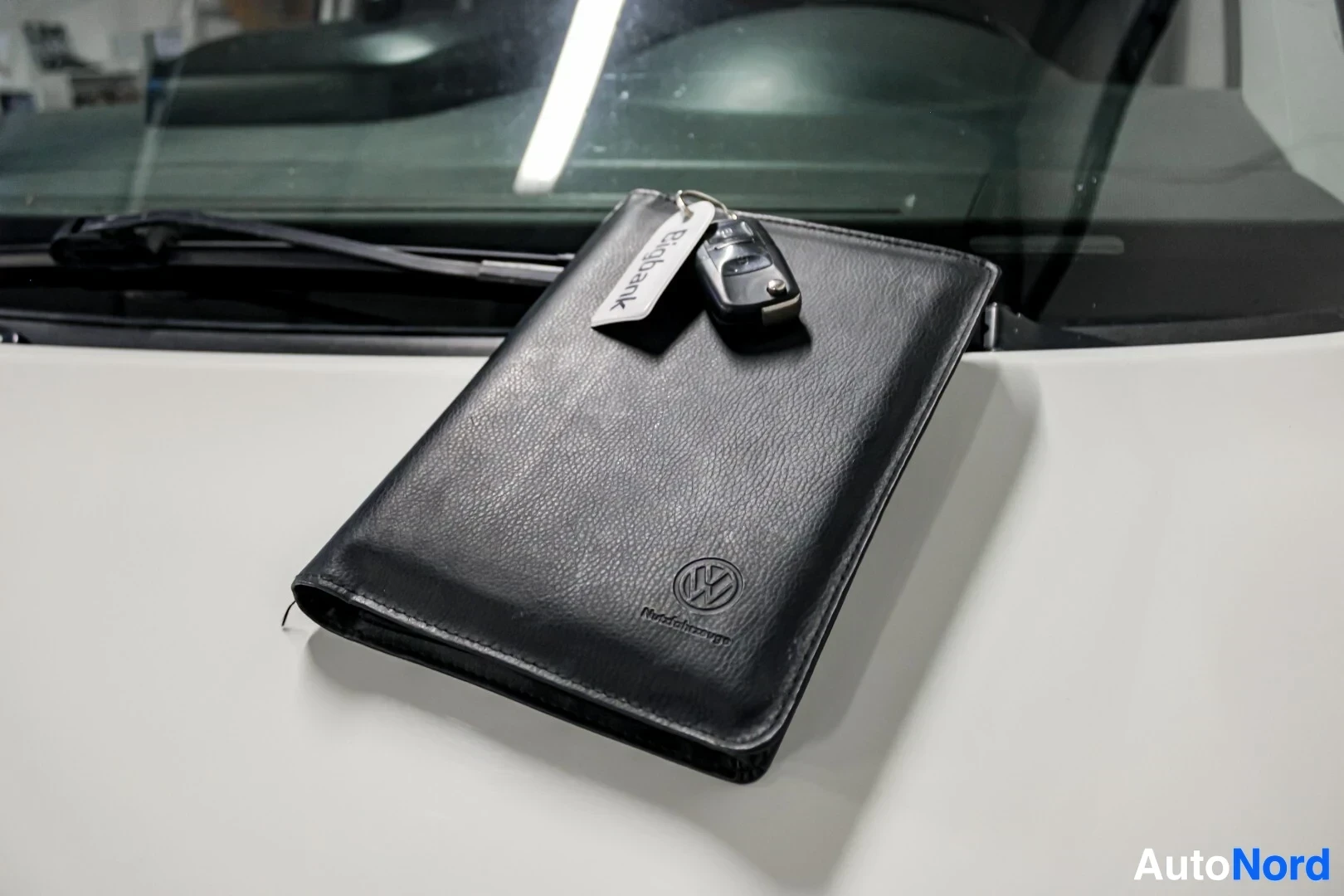 Yksityiskohta: Lähikuva mustasta nahkakansiosta, jossa on Volkswagen-logo, ja avaimesta avaimenperällä, jossa lukee "Bigbank". Esineet lepäävät valkoisen auton konepellillä tu