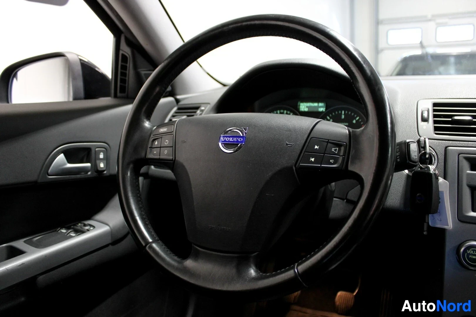 Ohjaamo: Ohjauspyörä edestäpäin nähtynä, jossa on Volvo-logo keskellä ja painikkeita molemmilla puolilla. Kojelauta ja osittain oven sisäpaneeli näkyvät taustalla. Avaim