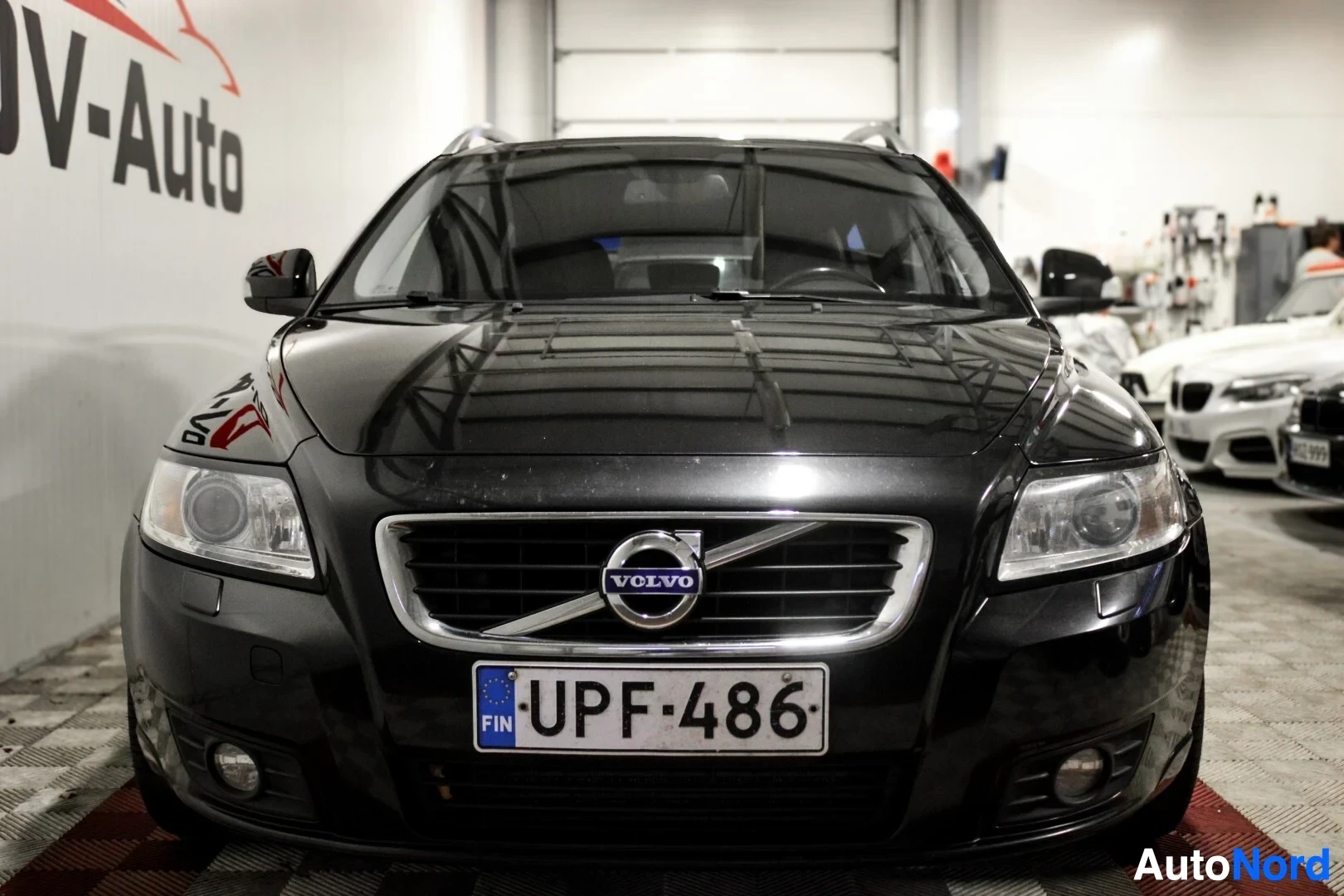 Edestä: Mustan farmariauton etuosa, jossa on Volvo-logo ja rekisterikilpi. Auto on pysäköity ruudulliselle lattialle sisätiloissa.