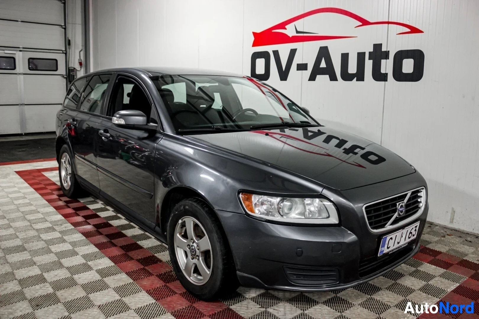 Etuviisto, nokka vasemmalle: Tummanharmaa Volvo V50 -farmariauto sisätiloissa kuvattuna viistosti edestä oikealta. Autossa on viisipuolaiset kevytmetallivanteet ja edessä suorakulm