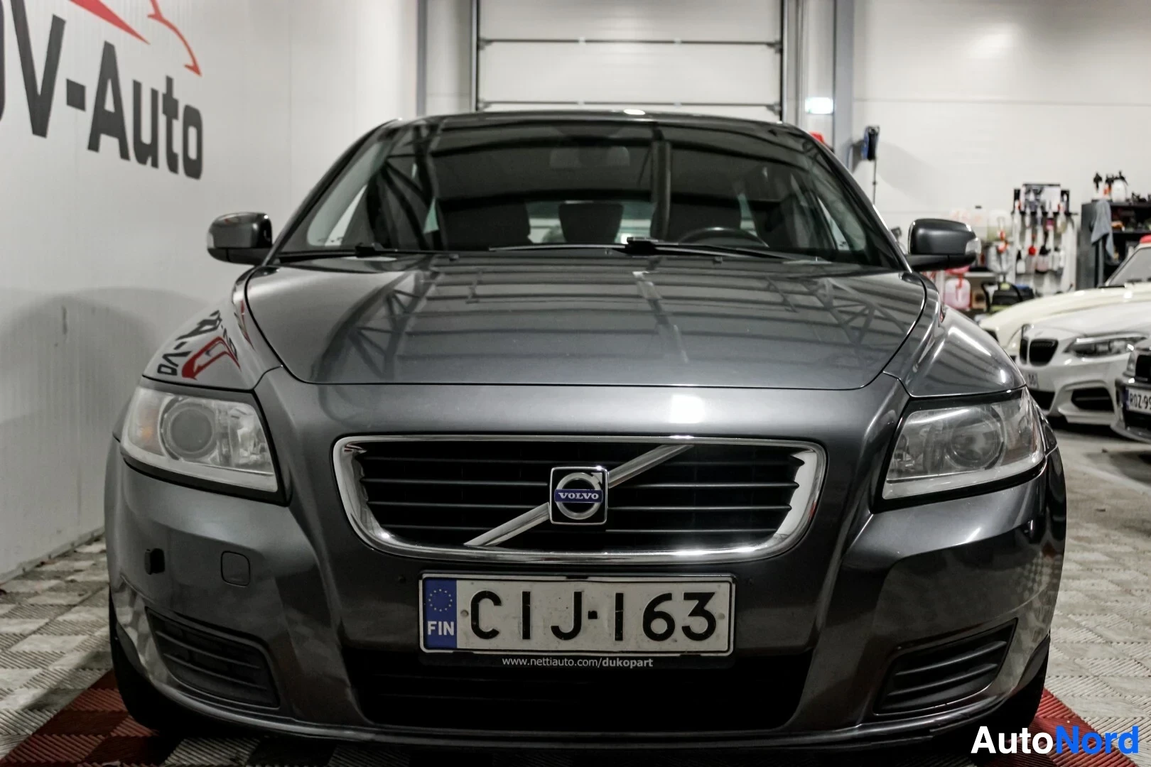 Edestä: Tummanharmaa Volvo V50 -farmariauto edestä päin kuvattuna sisätiloissa. Autossa on suorakulmaiset ajovalot ja etupuskurissa on parkkitutka-anturi.