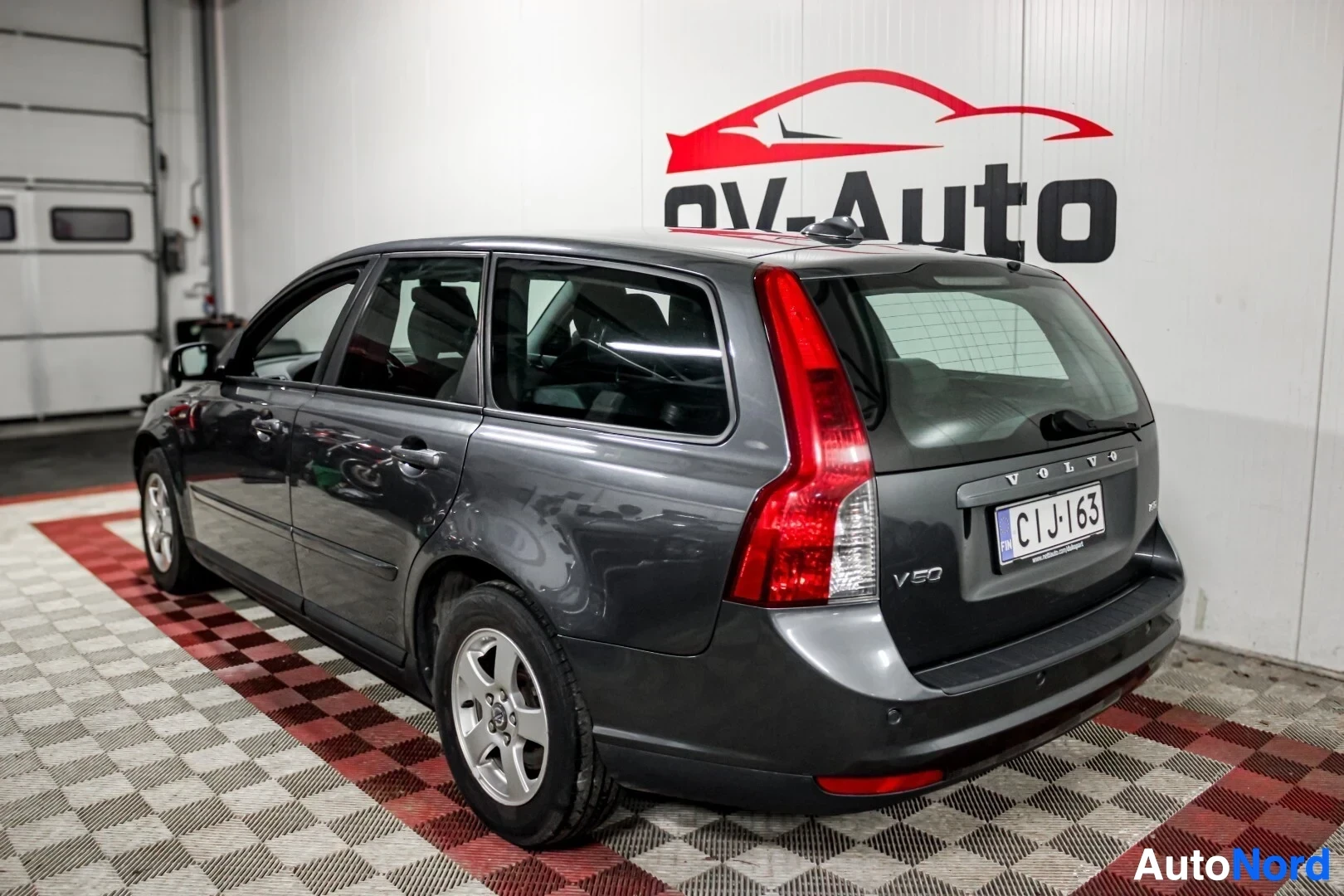 Takaviisto: Tummanharmaa Volvo V50 -farmariauto sisätiloissa kuvattuna viistosti takaa vasemmalta. Autossa on viisipuolaiset kevytmetallivanteet ja takapuskurissa on parkki
