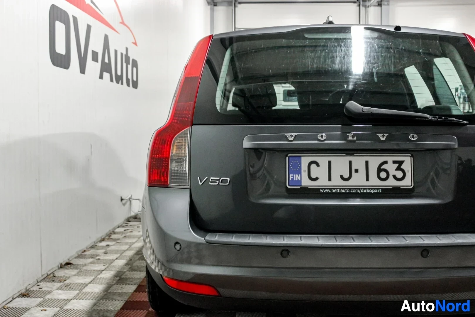 Takaa: Lähikuva harmaan farmariauton perästä. Takaluukussa on teksti "VOLVO", "V50" ja rekisterikilpi ""