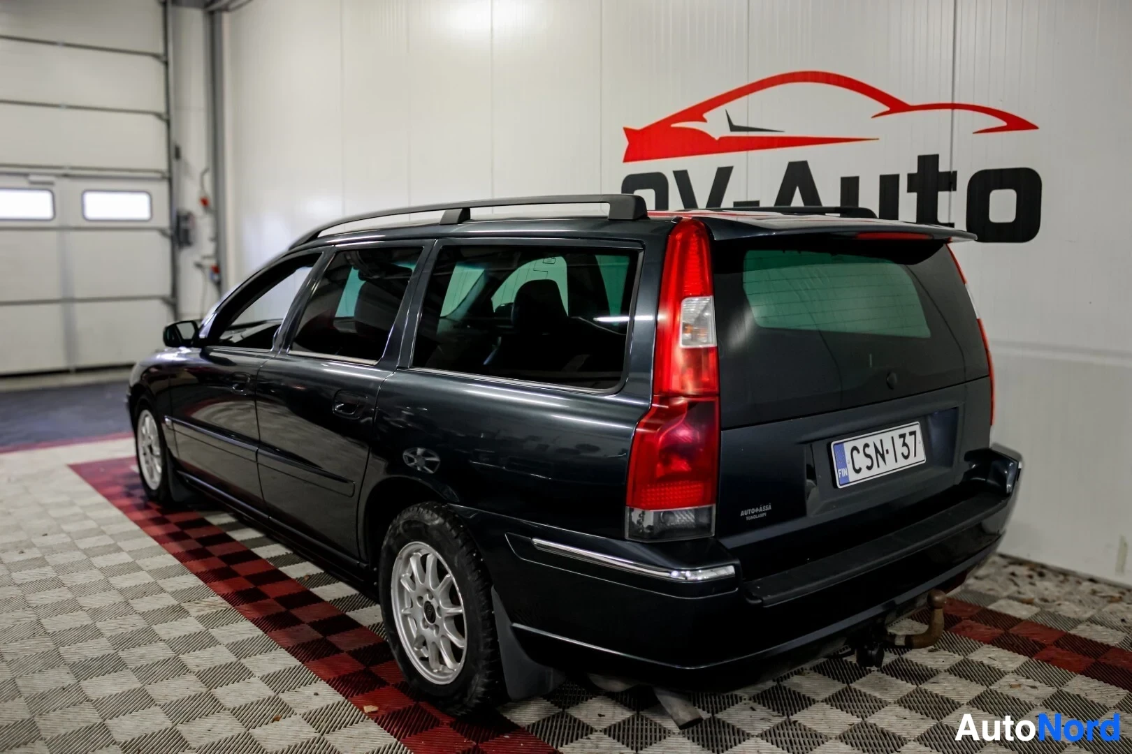 Takaviisto: Tummanharmaa farmariauto, jossa on kattokaiteet ja hopeanväriset vanteet, kuvattuna takaa oikealta. Takana näkyy vetokoukku ja rekisterikilpi ""