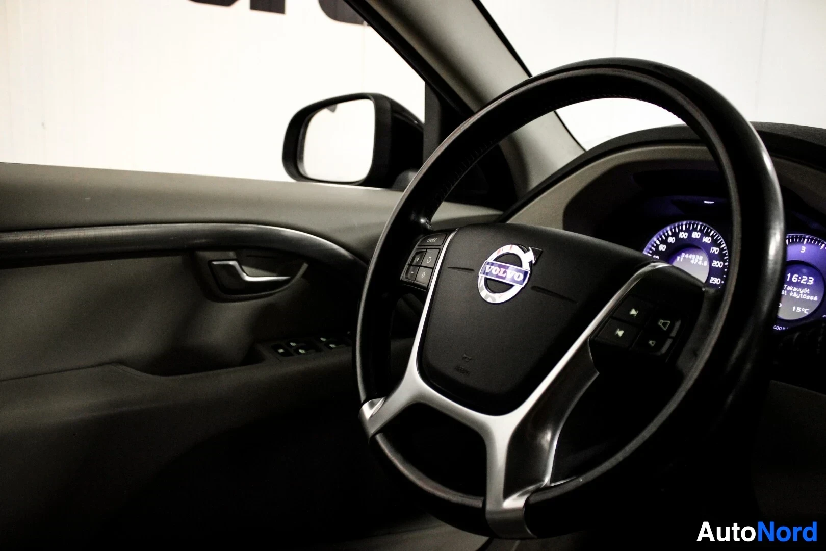 Ohjaamo: Lähikuva ohjauspyörästä, jossa on Volvo-logo ja monitoimipainikkeet. Taustalla näkyy auton oven sisäpaneeli.