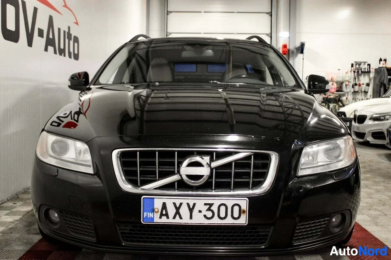 Edestä: Musta Volvo V70 -farmariauto edestä päin kuvattuna. Autossa on suorakaiteen muotoiset ajovalot ja kromireunainen etusäleikkö. Etupuskurissa on rekisterikilpi 'A