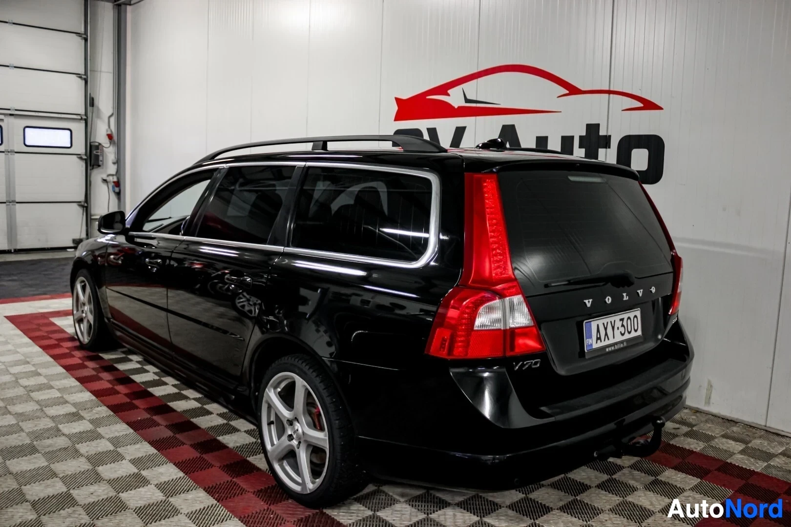 Takaviisto: Musta Volvo V70 -farmariauto kuvattuna takaa vasemmalta viistosti. Autossa on pystysuorat takavalot, kattokiskot ja hopeiset kevytmetallivanteet. Ajoneuvon taka
