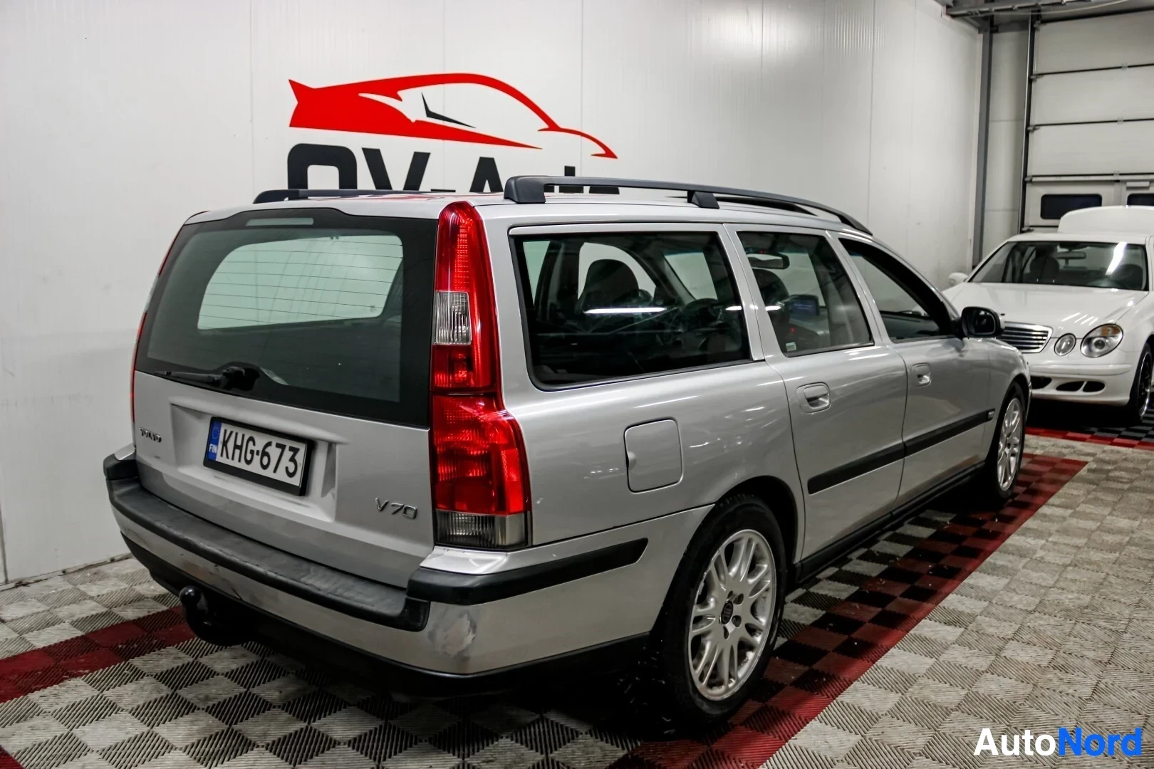Takaviisto: Hopeanvärinen farmariauto, Volvo V70, kuvattuna takavasemmalta viistosti. Ajoneuvossa on kattokiskot ja vetokoukku.