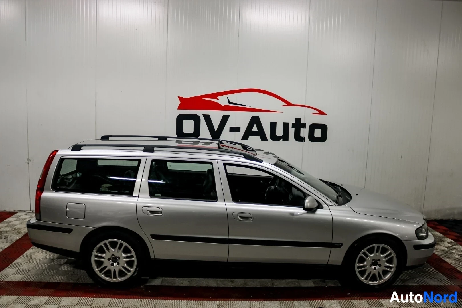 Sivulta: Hopeanvärinen Volvo V70 farmariauto kuvattuna oikealta sivulta. Autossa on kattokiskot ja kevytmetallivanteet.