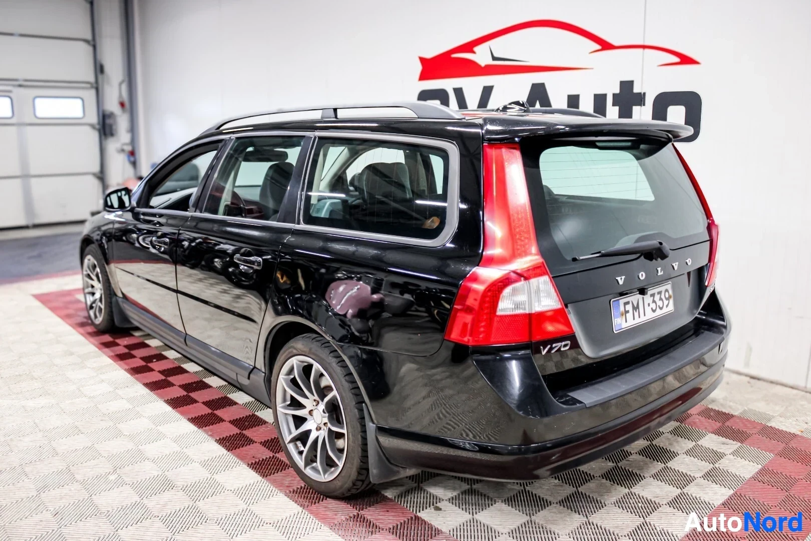 Takaviisto: Musta Volvo V70 -farmariauto kuvattuna takaviistosta oikealta sisätiloissa. Autossa on kattokaiteet ja takaluukussa on Volvo-logo, V70-merkki ja rekisterikilpi