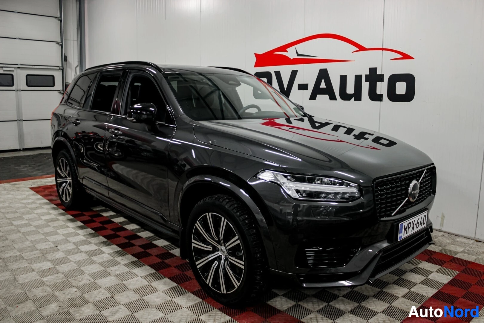Etuviisto, nokka oikealle: Tummanharmaa Volvo XC90, jossa on tummat monipuolaiset kevytmetallivanteet ja kattokaiteet, pysäköitynä esittelytilaan, jossa on OV-Auto-logo seinällä. A