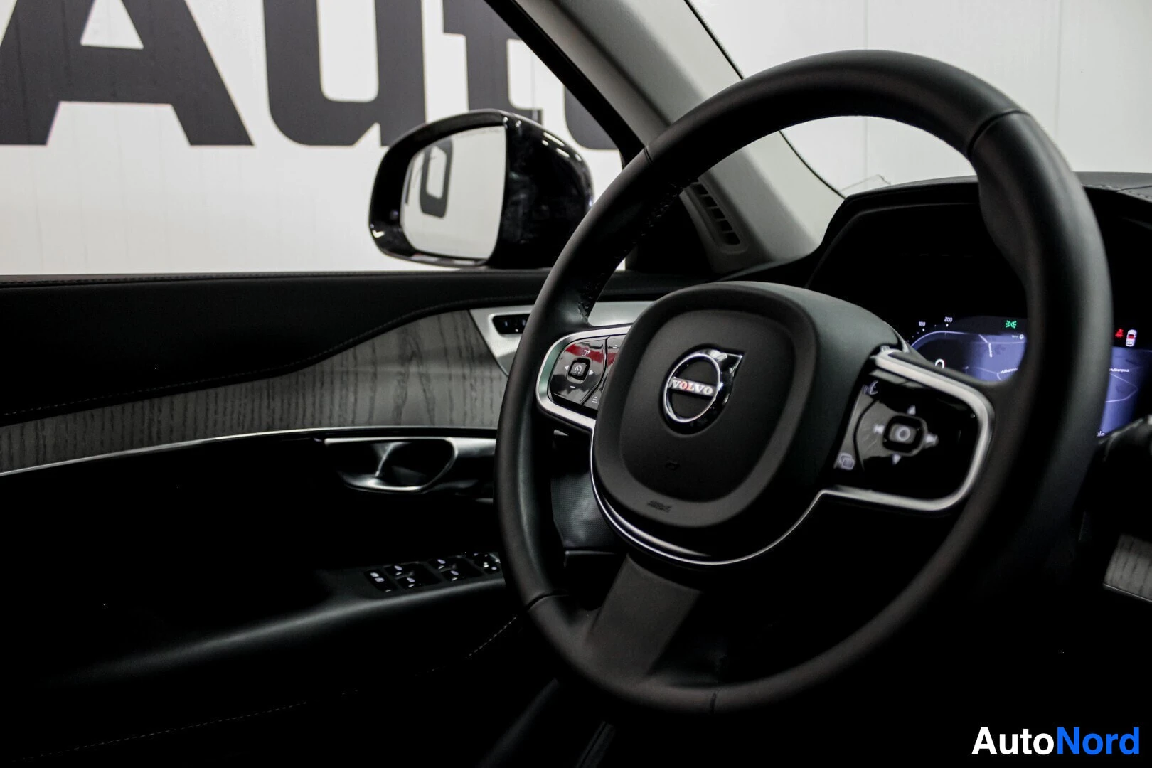Yksityiskohta: Musta nahkainen monitoimiohjauspyörä, jossa on Volvo-logo. Ohjauspyörän takana näkyy osa digitaalisesta mittaristosta ja vasemmalla oven sisäpaneeli, jossa on p