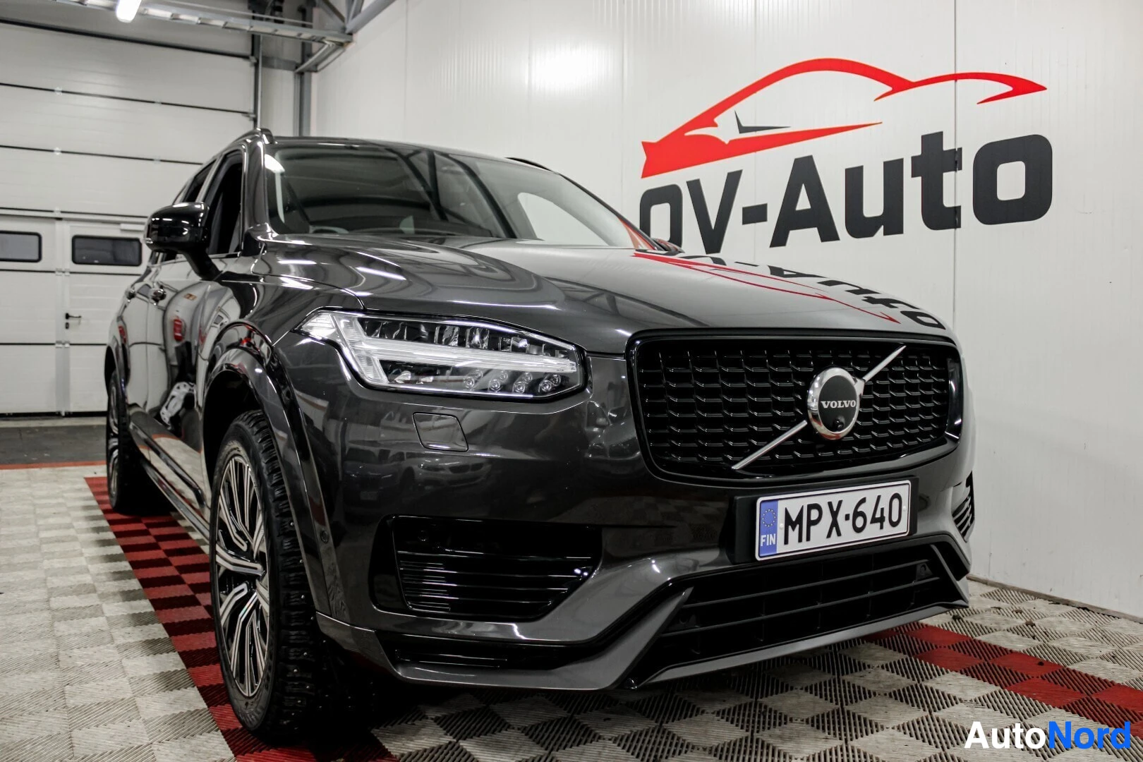 Etuviisto, nokka oikealle: Tummanharmaa Volvo XC90 edestä oikealta kolmen neljäsosan kuvakulmasta, esittelytilan valkoista seinää vasten. Autossa on LED-ajovalot ja rekisterikilpi