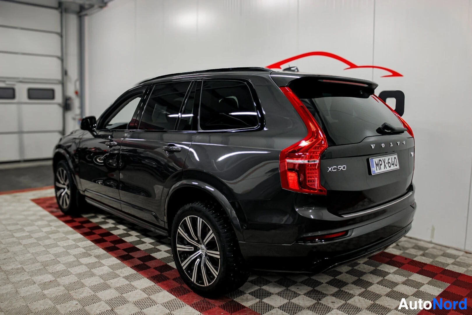 Takaviisto: Musta Volvo XC90, jossa on mustat talvirenkaat ja monipuolaiset kevytmetallivanteet, näkyy takaviistosta oikealta sisätilassa. Ajoneuvon rekisterikilpi on "MPX-