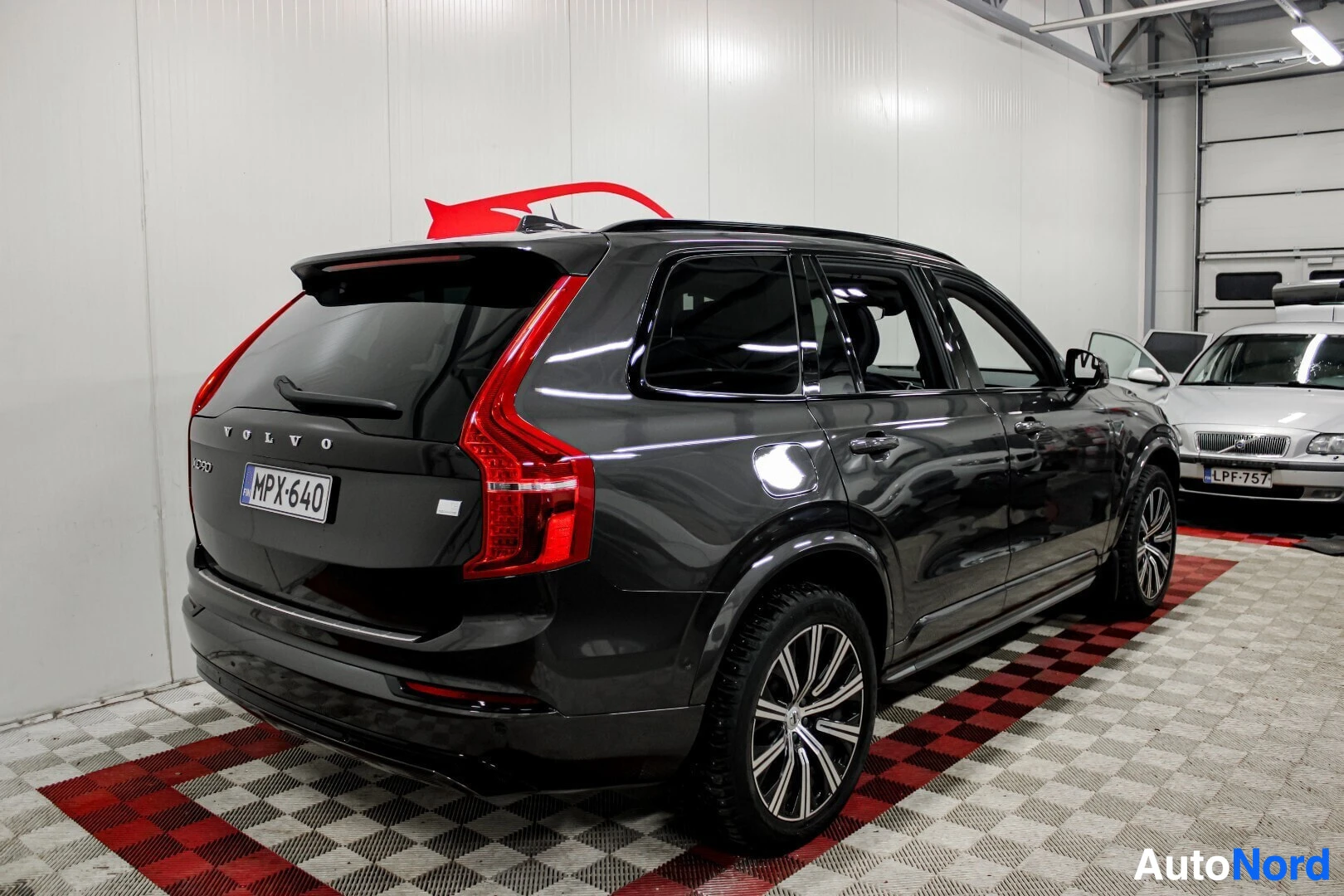 Takaviisto: Musta Volvo XC90, jossa on mustat talvirenkaat ja monipuoliset kevytmetallivanteet, näkyy takaviistosta vasemmalta sisätilassa. Ajoneuvon rekisterikilpi on "MPX