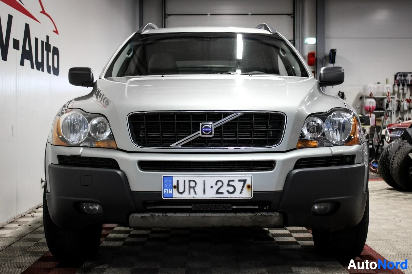 Edestä: Hopeanvärisen Volvo XC90 -auton etuosa on kuvattu suoraan edestä. Auton rekisterikilpi on "". Rekisterikilven yläpuolella näkyy Volvo-logo. Ajoneuvo on p