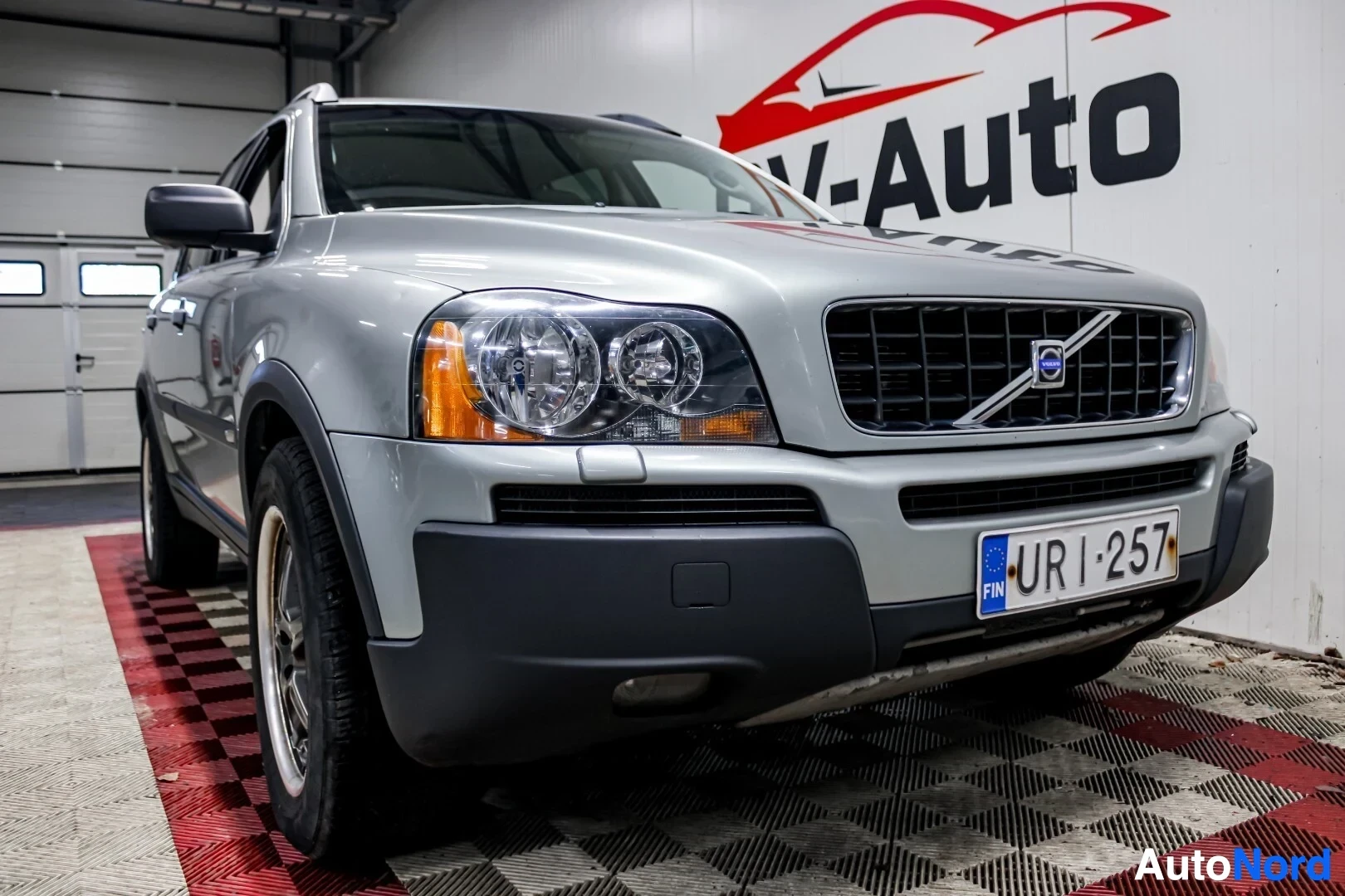 Etuviisto, nokka oikealle: Hopeanvärinen Volvo XC90 on kuvattu alhaalta ja edestä, keula osoittaa hieman oikealle. Auton rekisterikilpi on "". Takana näkyy OV-Auto-logo ja r