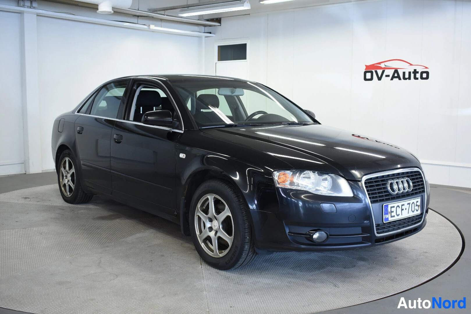 Etuviisto, nokka oikealle: Musta Audi A4 -henkilöauto kuvattuna etuvasemmalta viistosti. Auton rekisterikilvessä on teksti. Autossa on viisipuolaiset kevytmetallivanteet. T